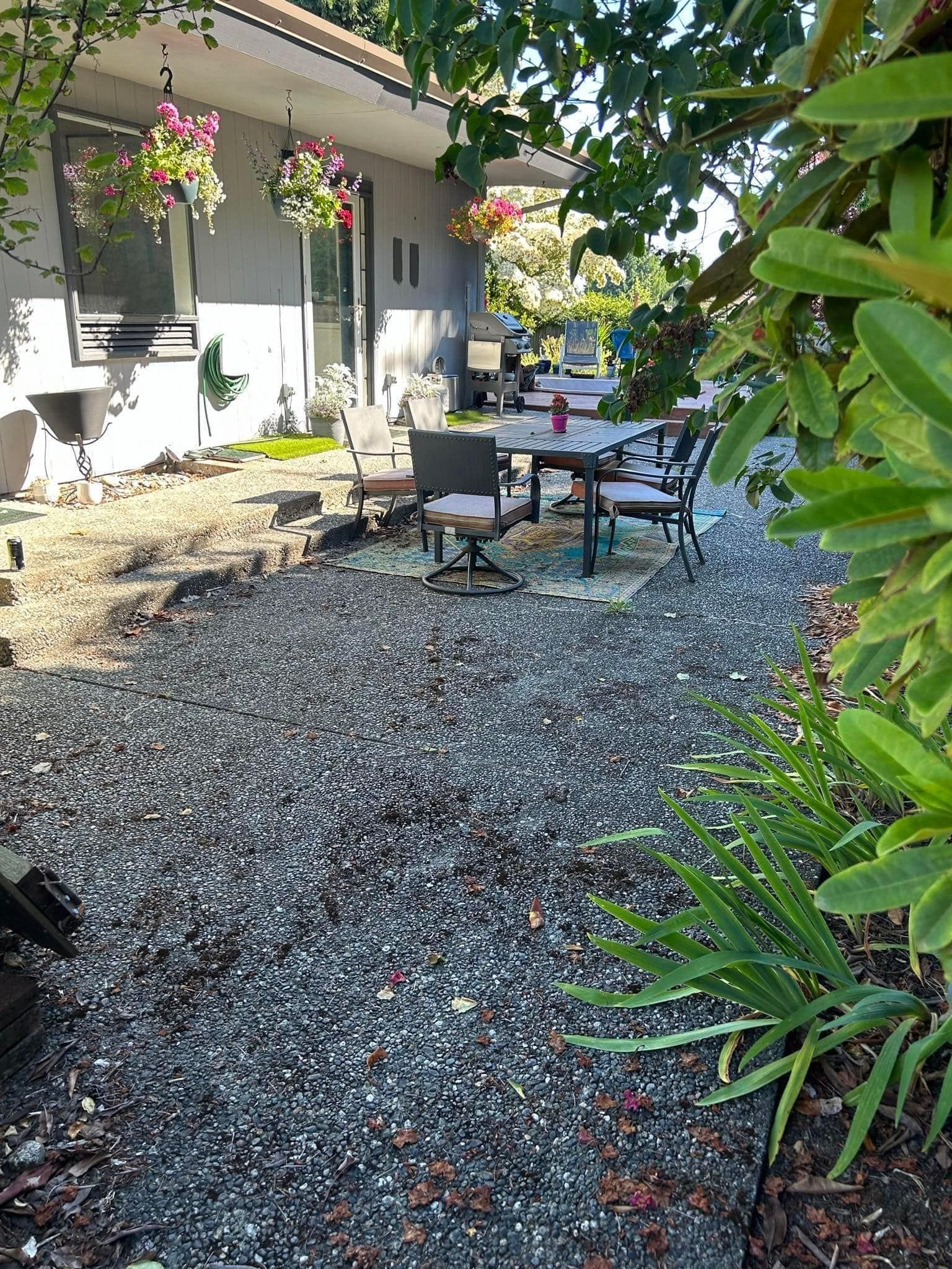 Gravel patio alongside a house with a table and chairs, surrounded by greenery.