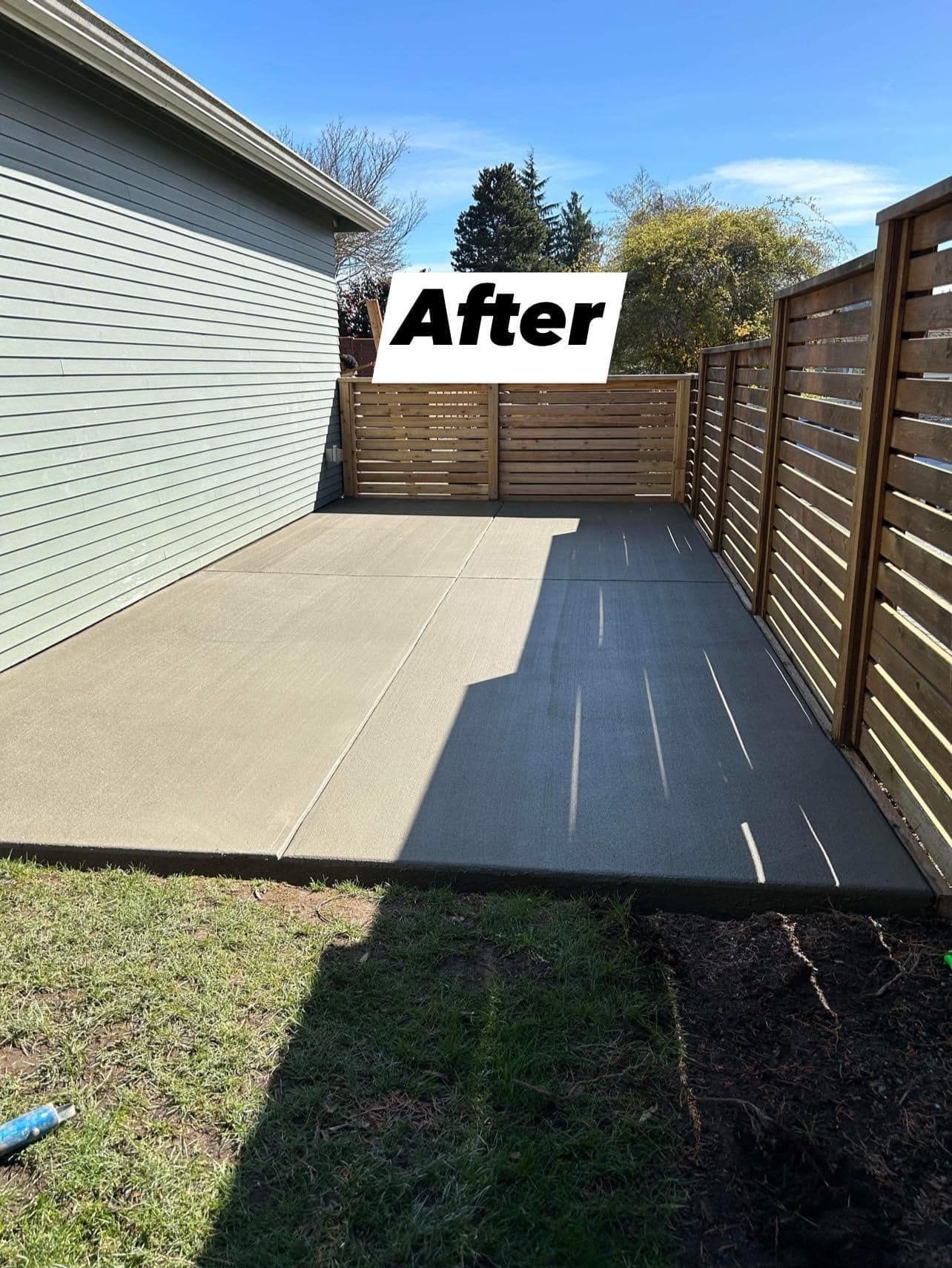 Newly poured concrete patio with wooden fence, next to a house under a blue sky.