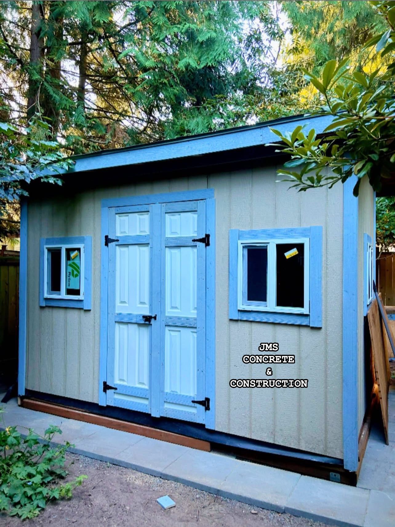 Tan shed with blue trim, double doors, and windows, set in a backyard.