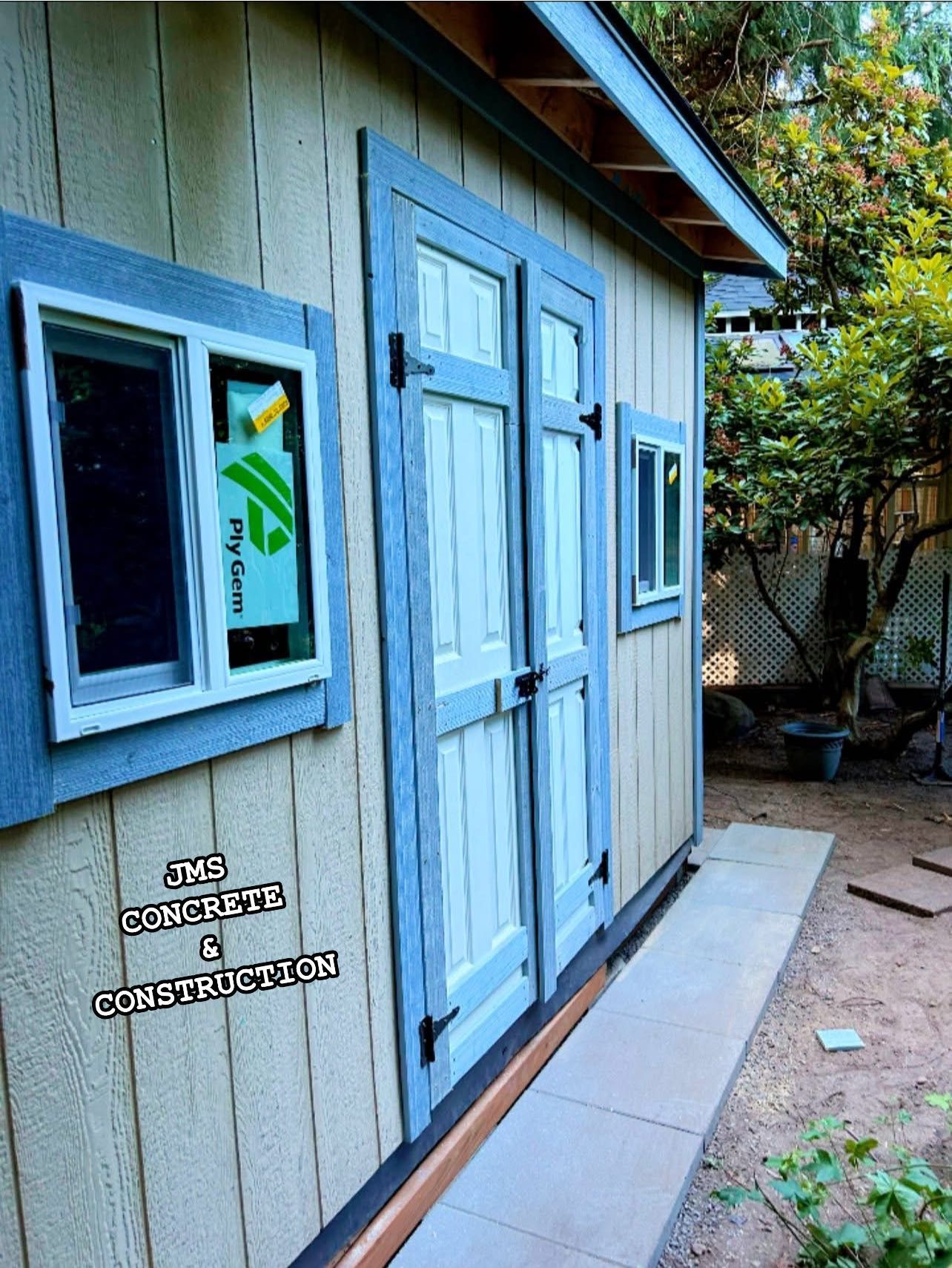A shed with blue-trimmed doors and windows, light-colored siding, and a concrete path.
