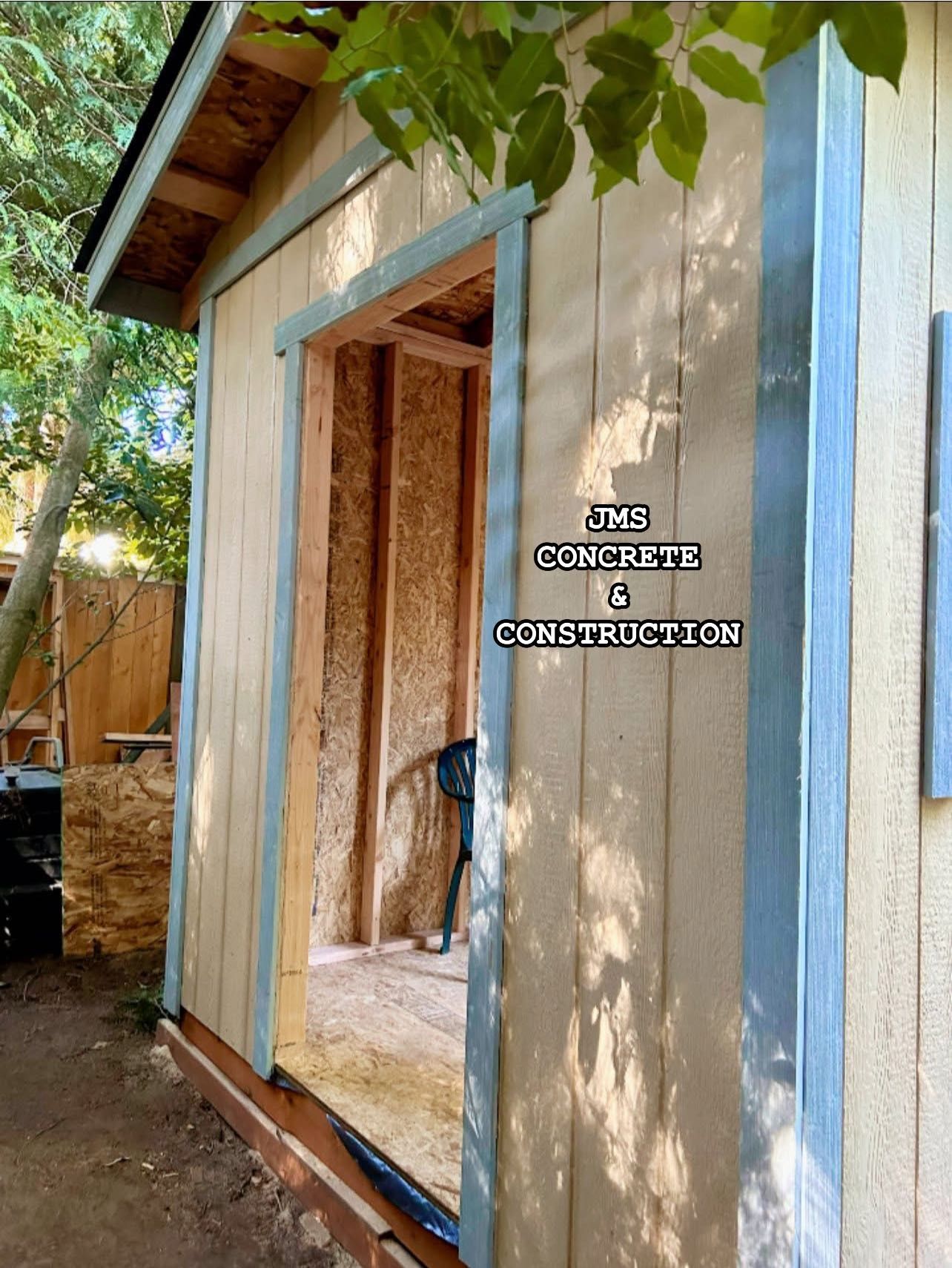 Wooden shed under construction, doorway open. Beige siding, blue trim. Backyard setting.