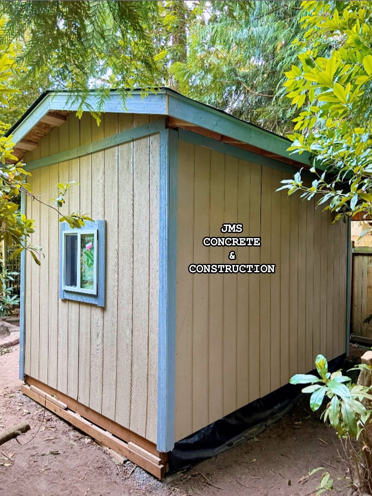 Tan shed with blue trim and a small window surrounded by greenery.