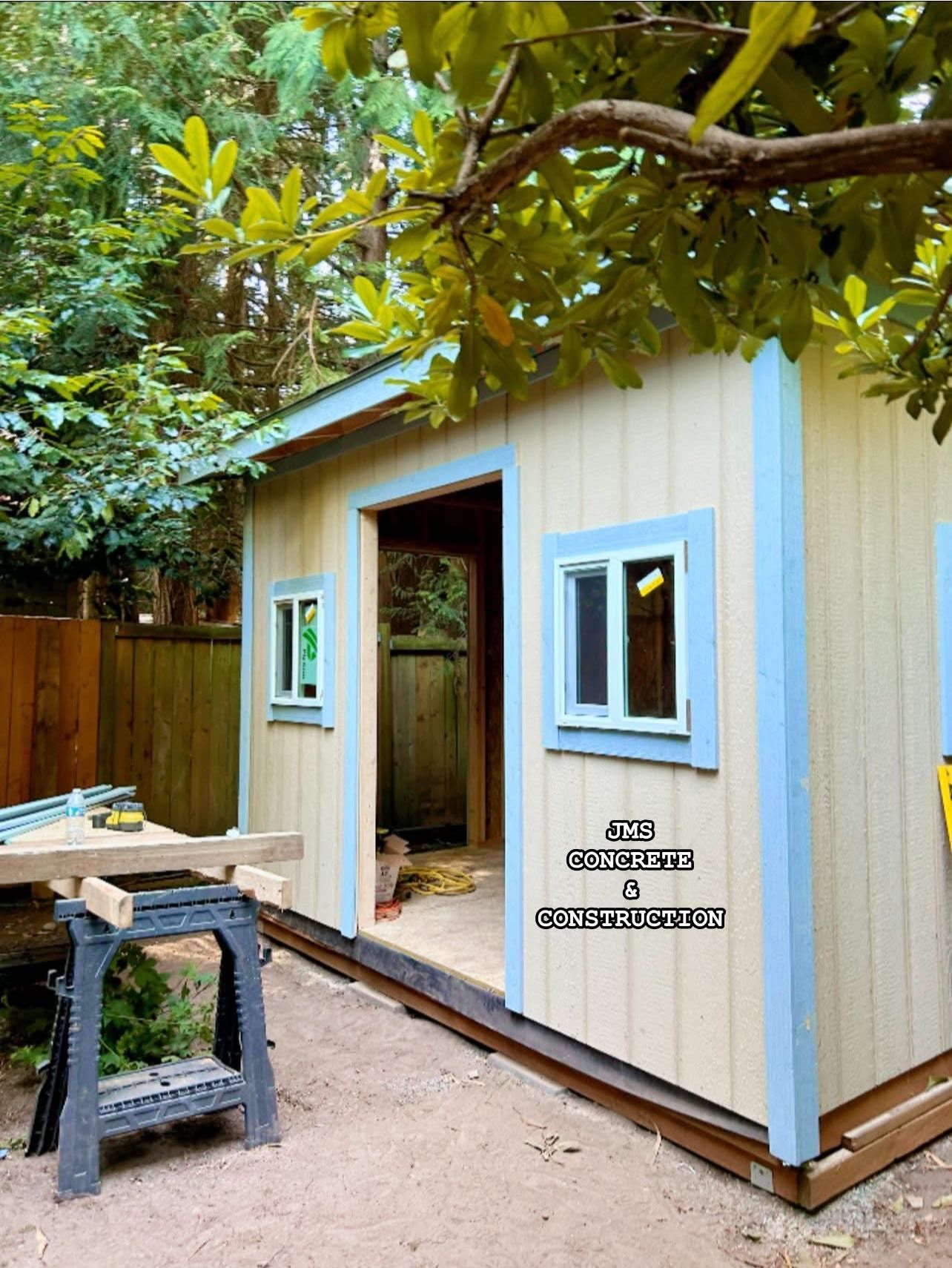Beige shed with blue trim and two windows under tree branches; construction in progress.