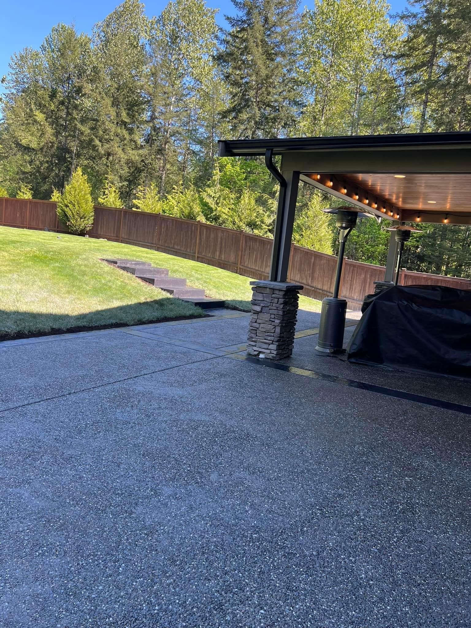 Gravel patio next to a grassy lawn and a covered patio with a black cover. Wooden fence and trees in the background.