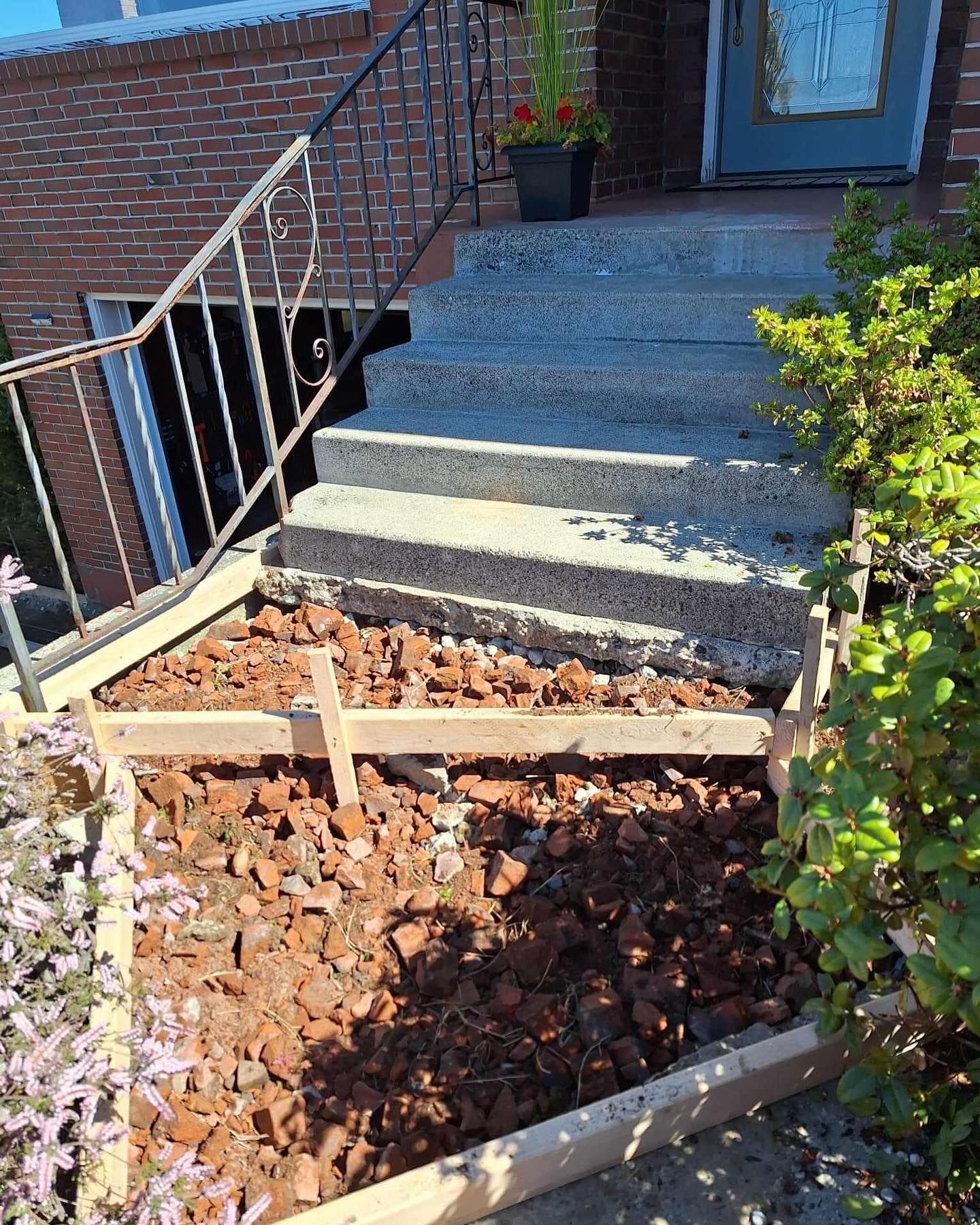 Concrete steps leading up to a house with a damaged lower section undergoing repair; reddish-brown gravel visible.