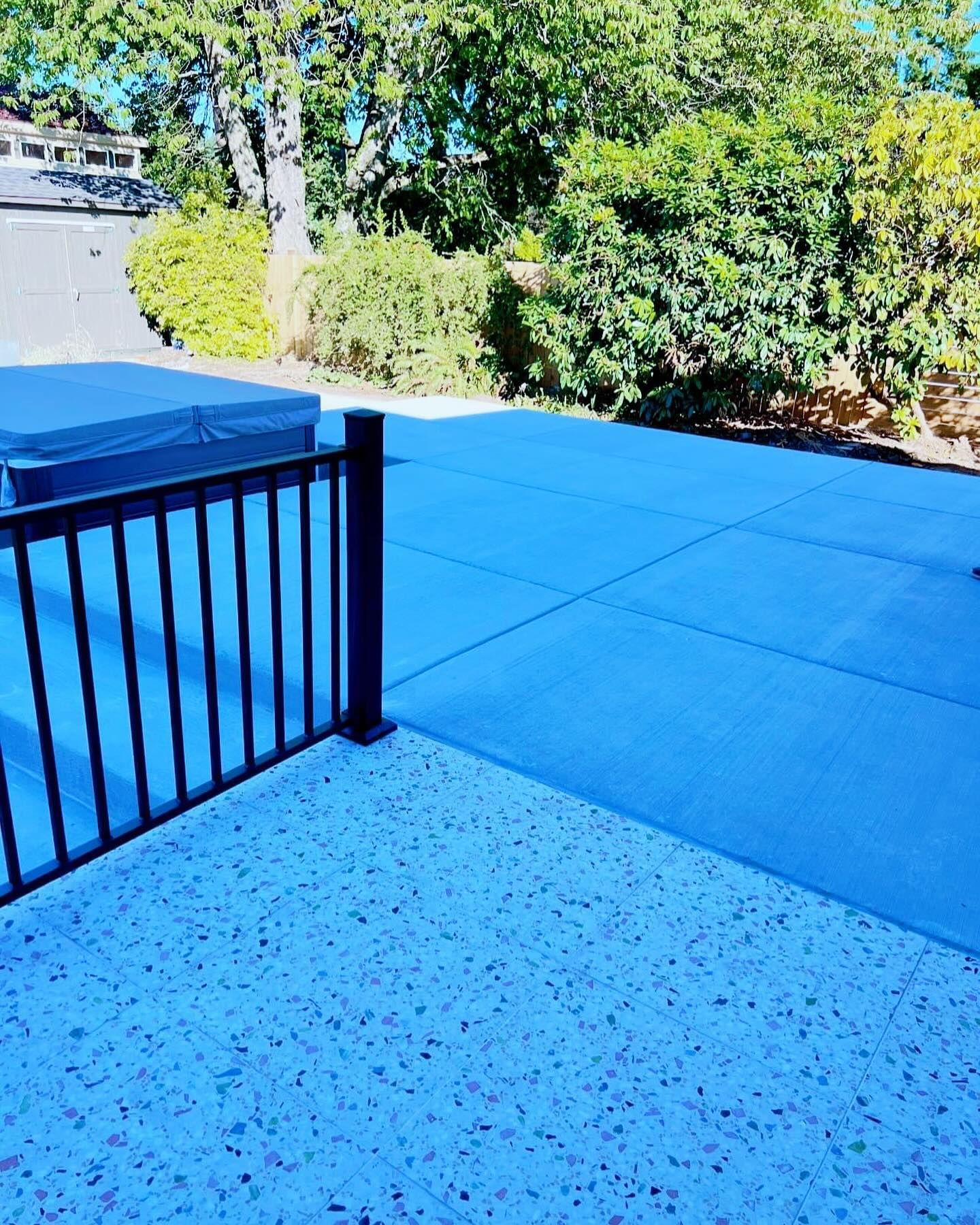 Patio with black railing and concrete squares, hot tub in the background, green trees, overcast day.