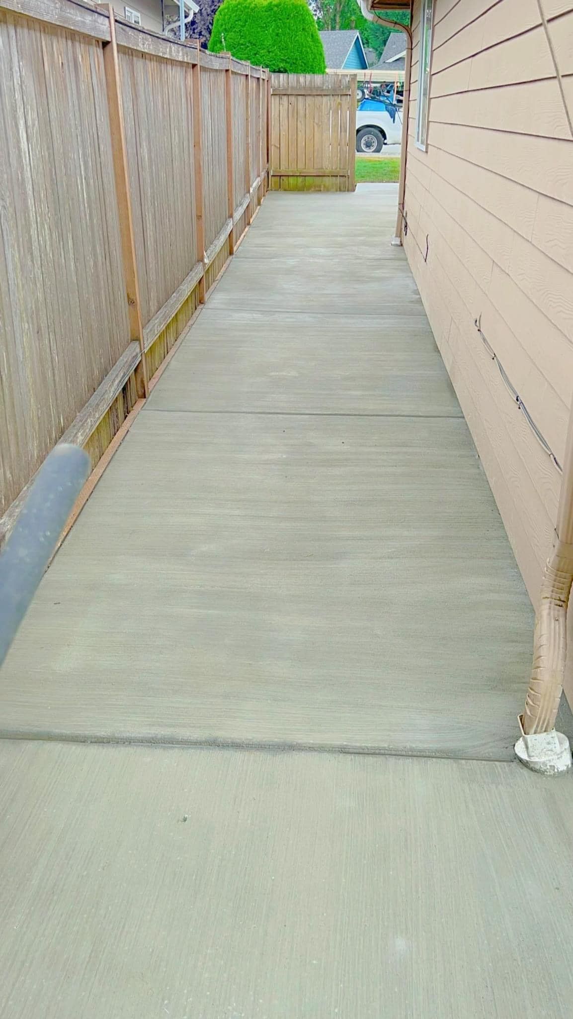 Narrow concrete walkway between a wooden fence and a house with beige siding.
