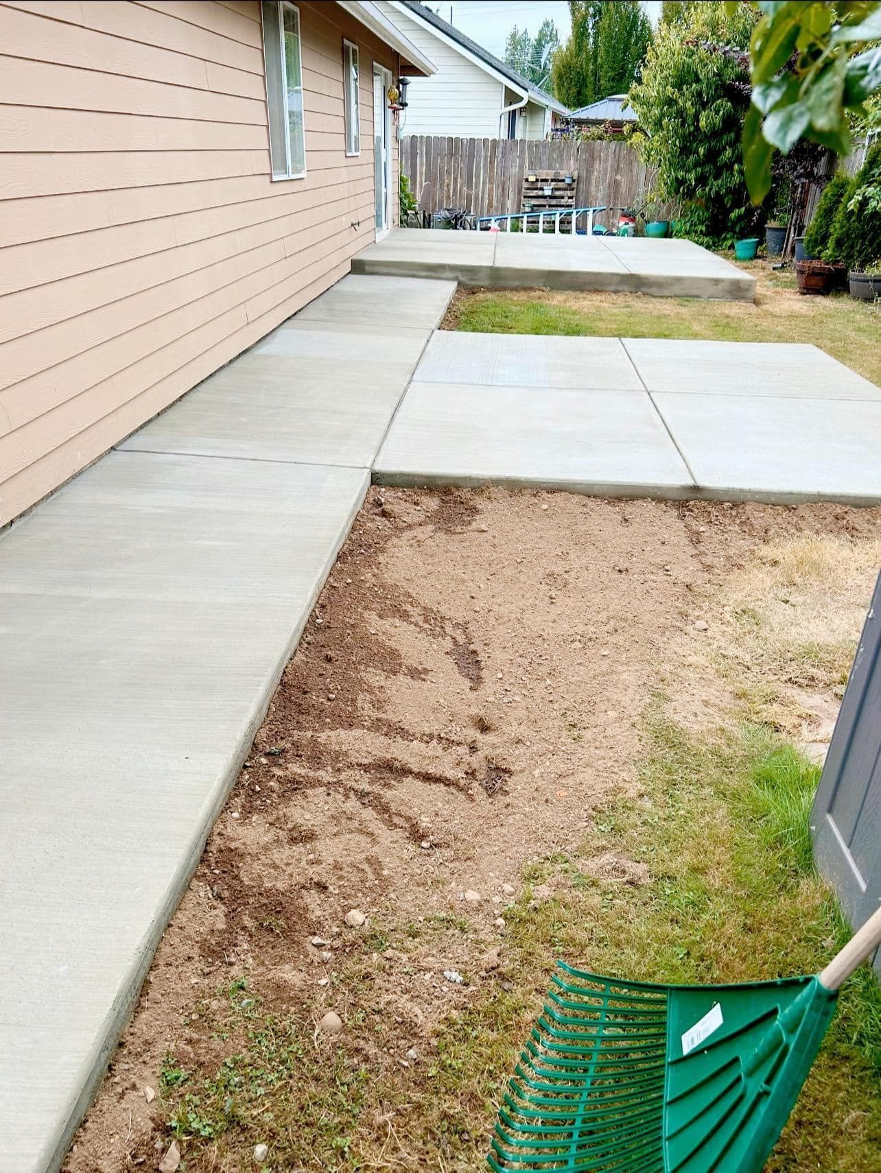 Concrete patio and walkway alongside a tan house. Bare dirt and a green rake in the foreground.