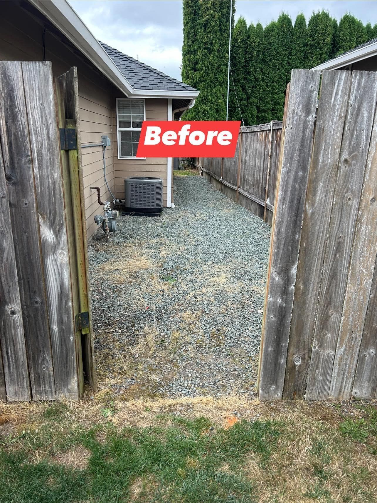 Open wooden gate revealing gravel path between two fences, leading to a house with an air conditioner.