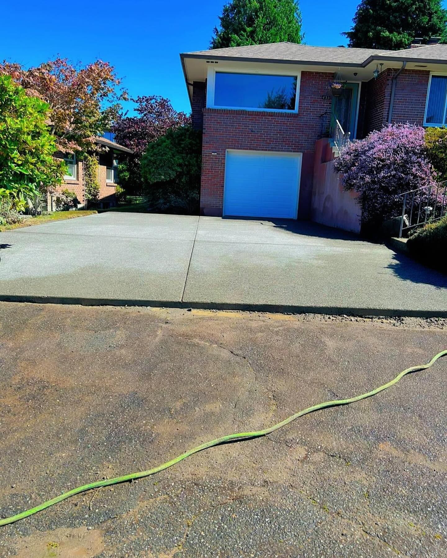 Driveway leading to a brick house with a garage, green hose, and nearby trees under a blue sky.