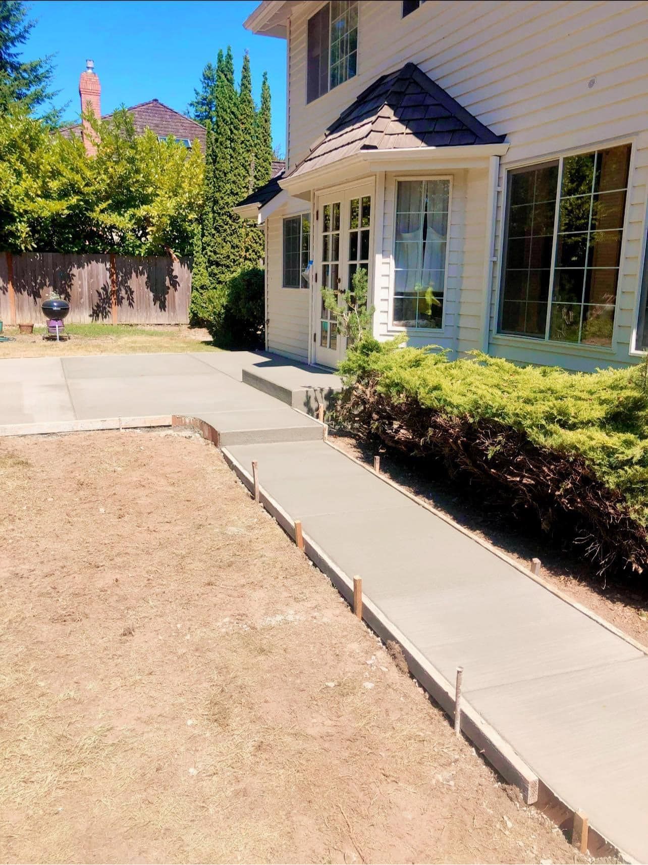A freshly poured concrete walkway leads to a light-colored house. Wooden frames border the new path, set on dry grass.