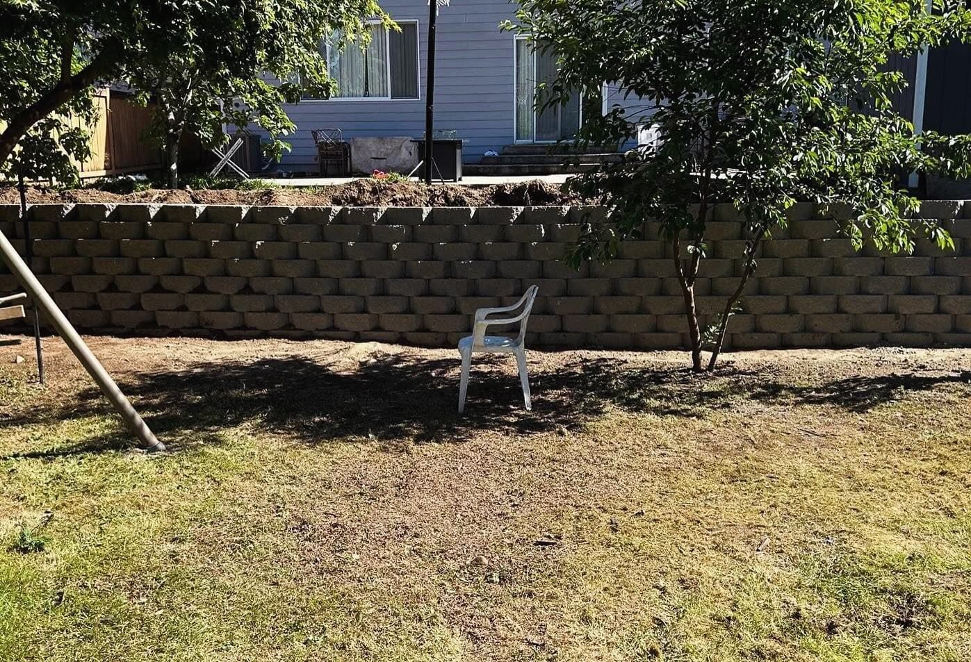 White chair in a grassy yard, in front of a retaining wall. Trees cast shadows. A light blue house is in the background.