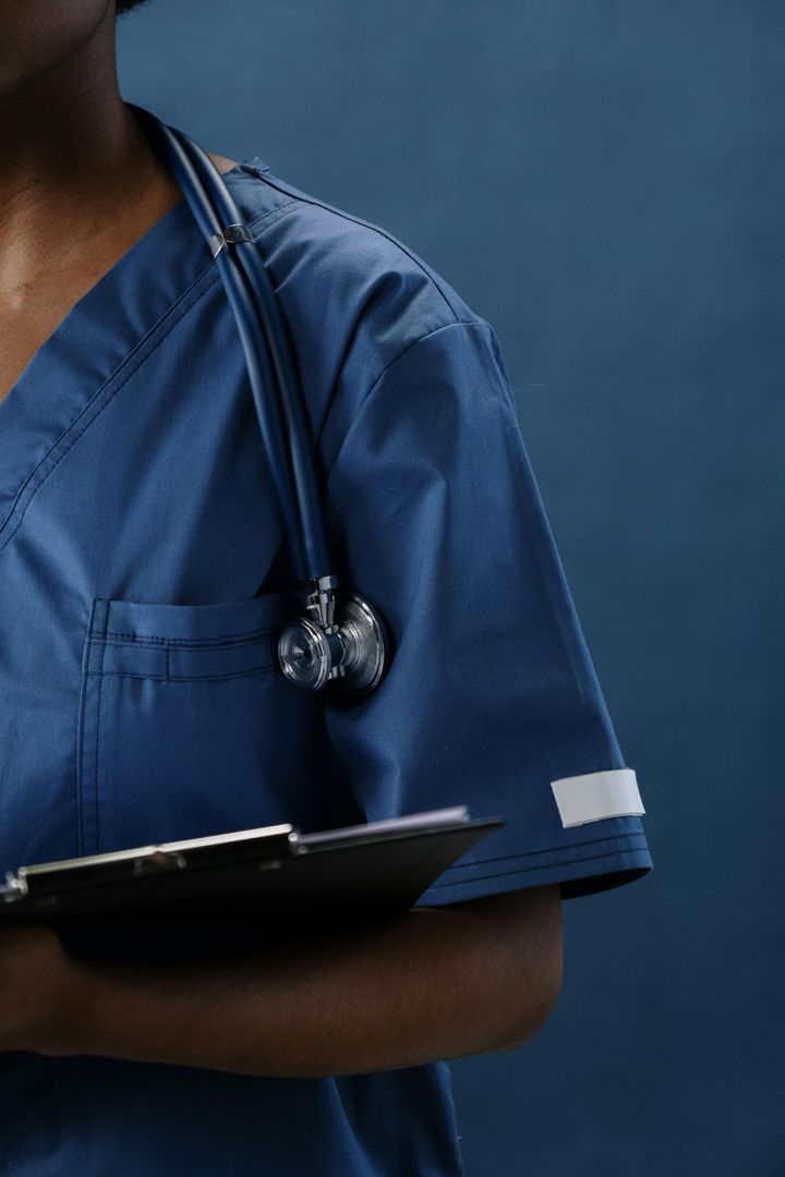 A person in blue scrubs with a stethoscope around their neck holds a clipboard.