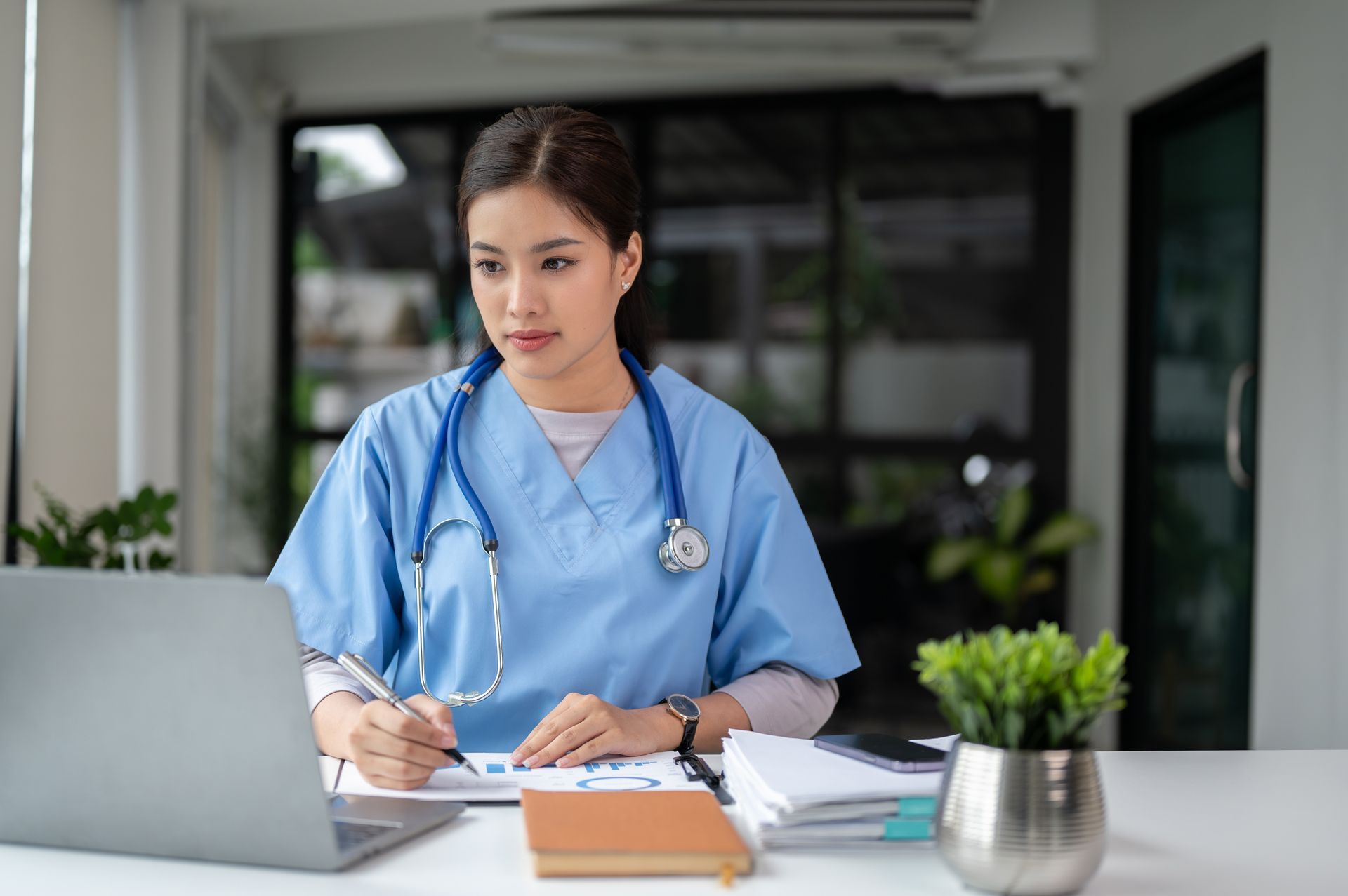 Medical professional in blue scrubs using a laptop and writing at a desk with a stethoscope.