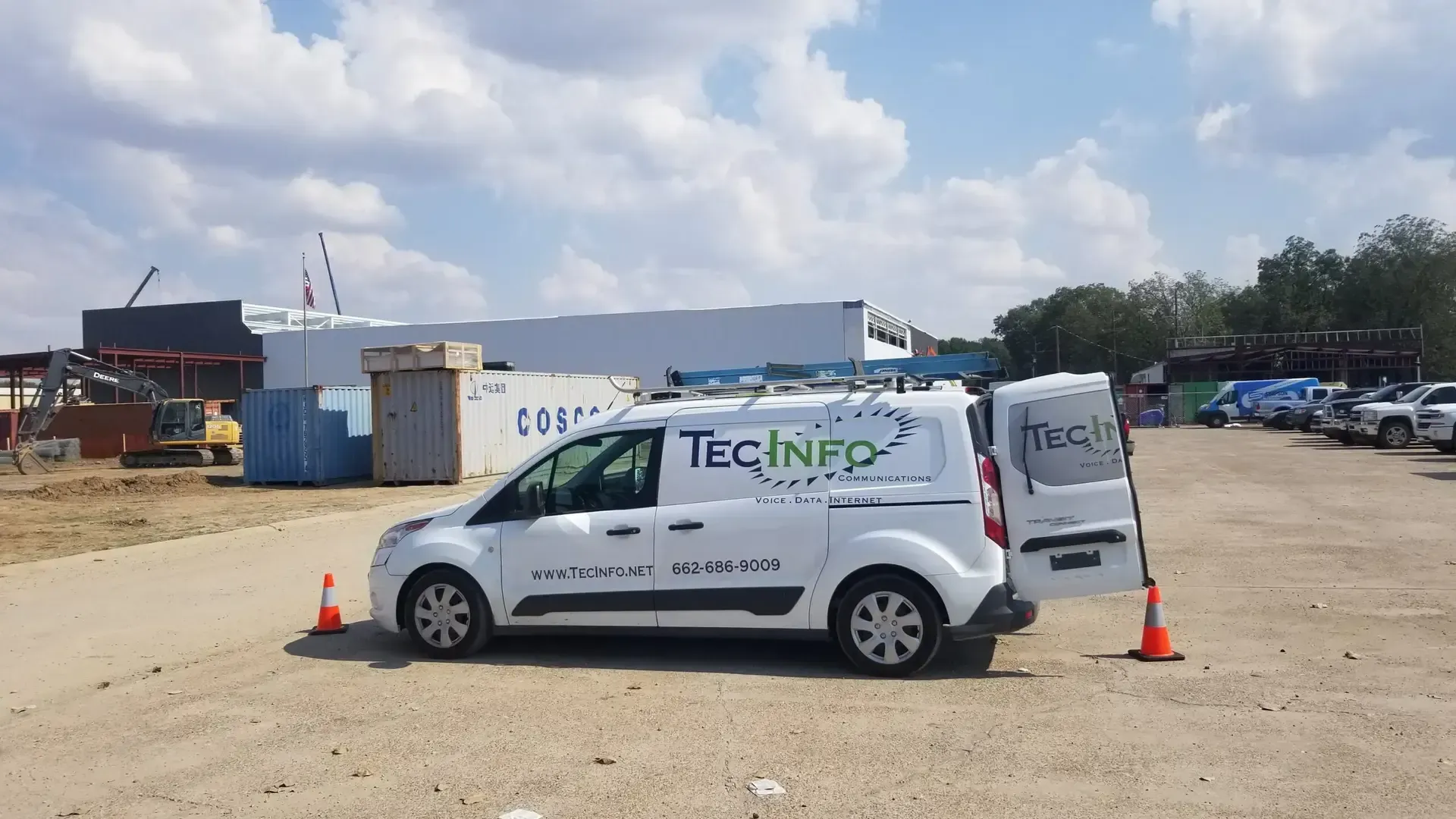 White TECINFO van parked on a gravel lot with orange cones, industrial buildings in the background.