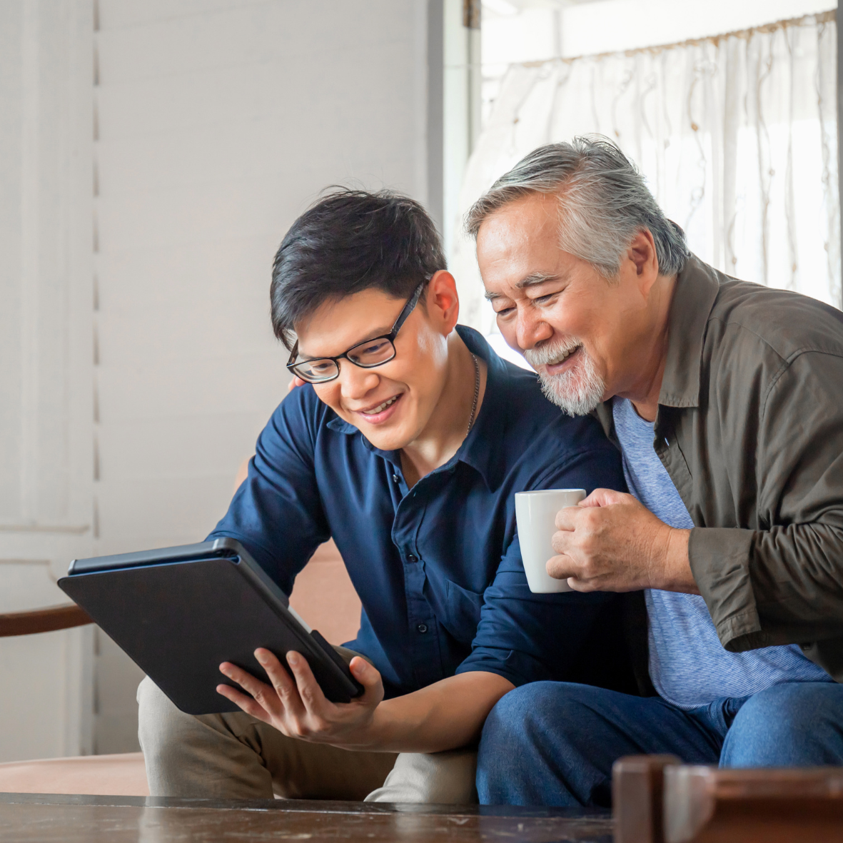 Two people smiling while looking at a tablet and holding a mug on a couch