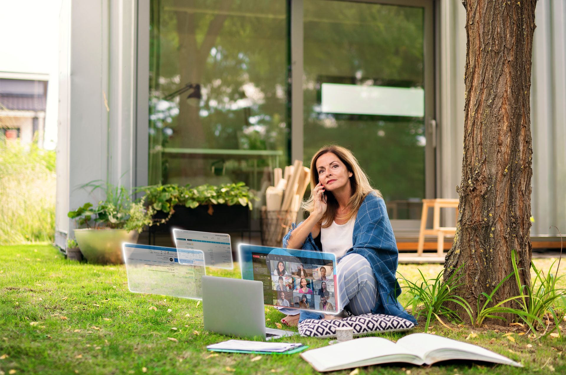 Woman sitting on grass with a laptop and notebooks near a tree outside a modern building