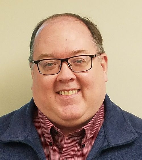Smiling man wearing glasses, red shirt, and blue jacket against a plain beige background