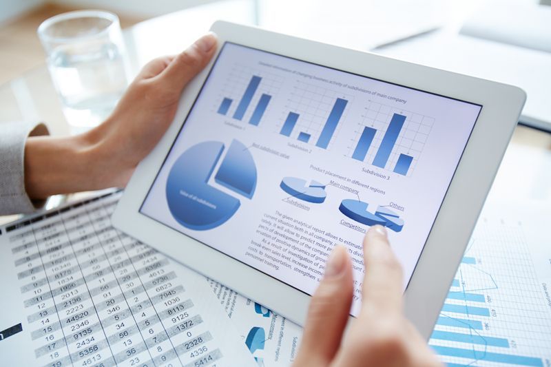 Hands holding a tablet displaying blue business charts and graphs over office paperwork