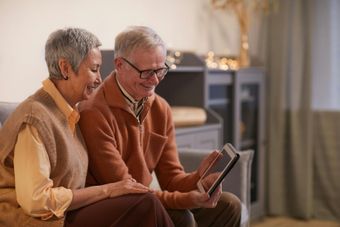 Two older adults sitting together, smiling and looking at a tablet in a cozy living room