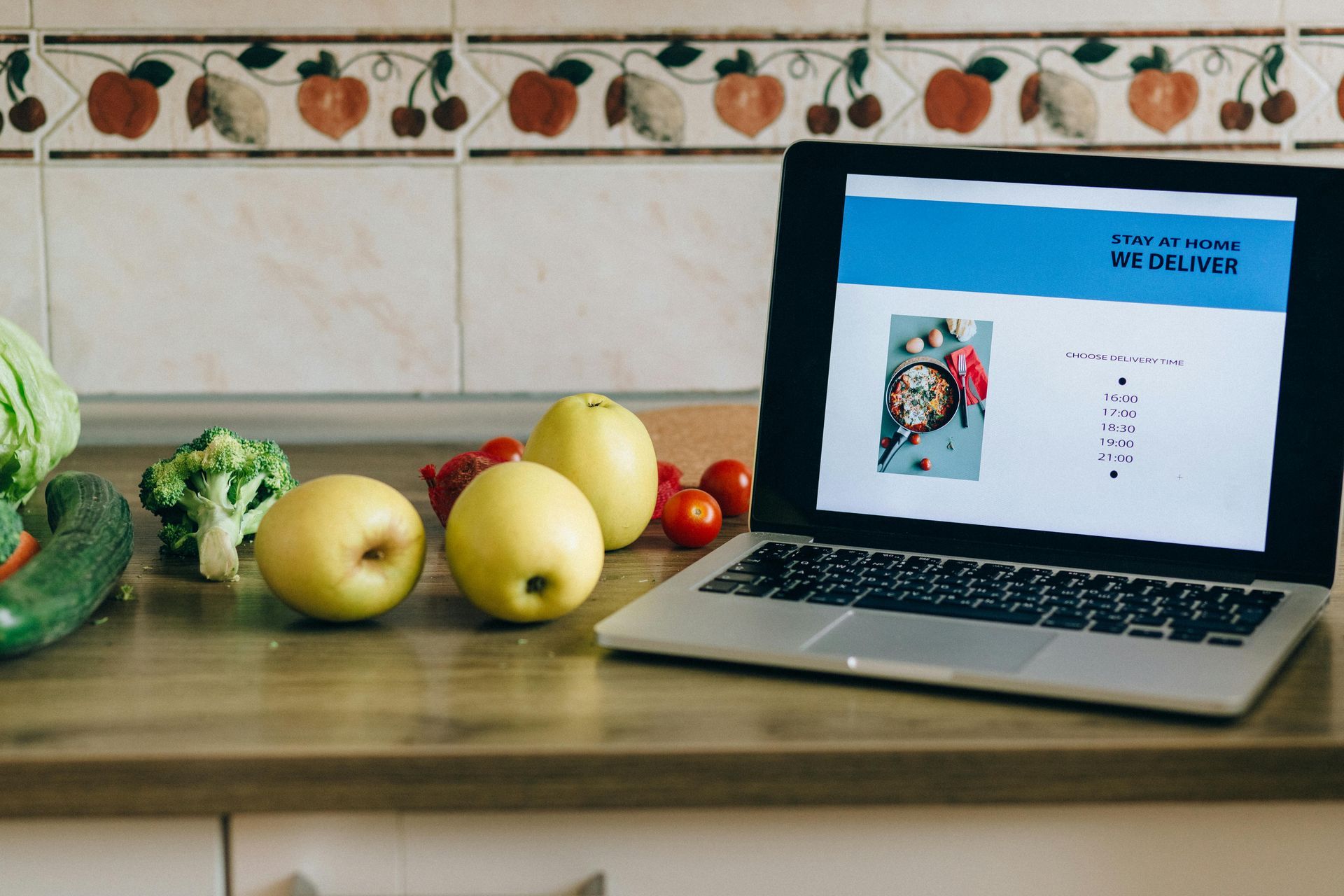 Laptop on a kitchen counter beside apples, broccoli, tomatoes, and a blue-screen page.