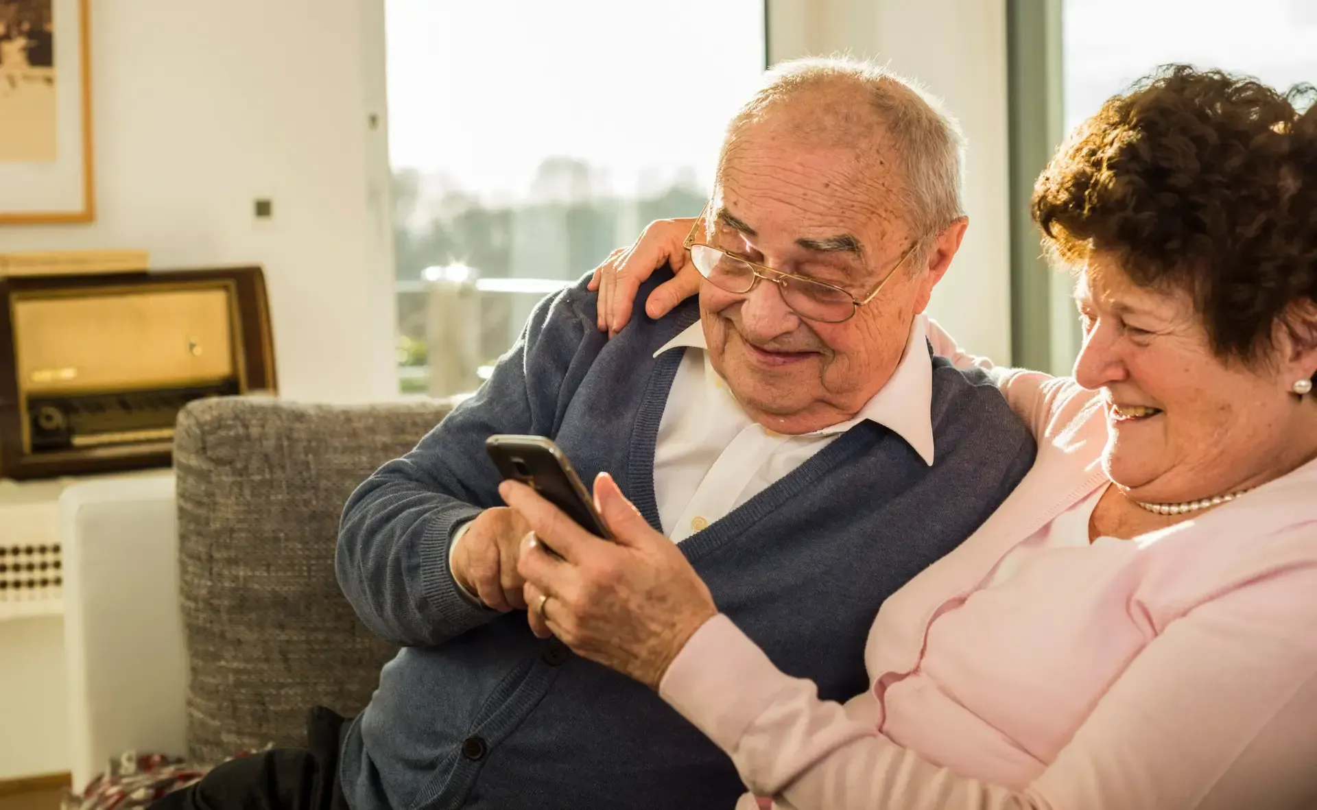 Two people smiling on a couch, looking at a smartphone together in a bright living room