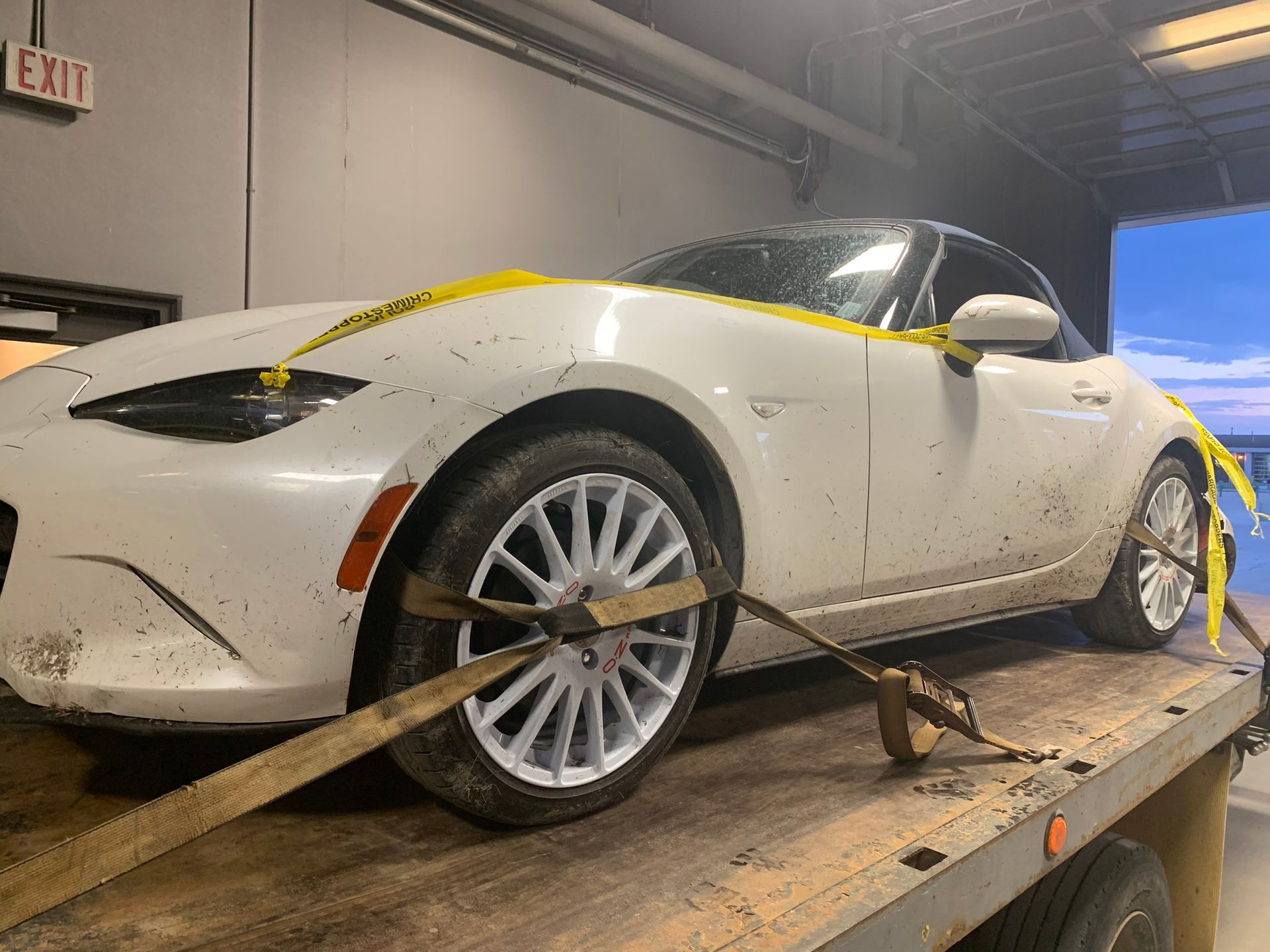White sports car strapped to a trailer in a warehouse, with yellow tape across the body.