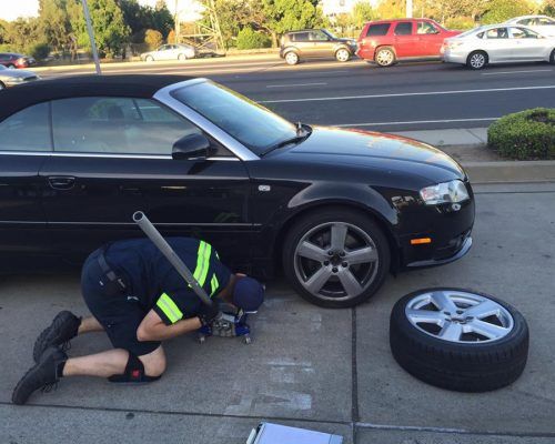 Mechanic kneels beside a black car, changing a flat tire on a roadside, with the wheel removed nearby.