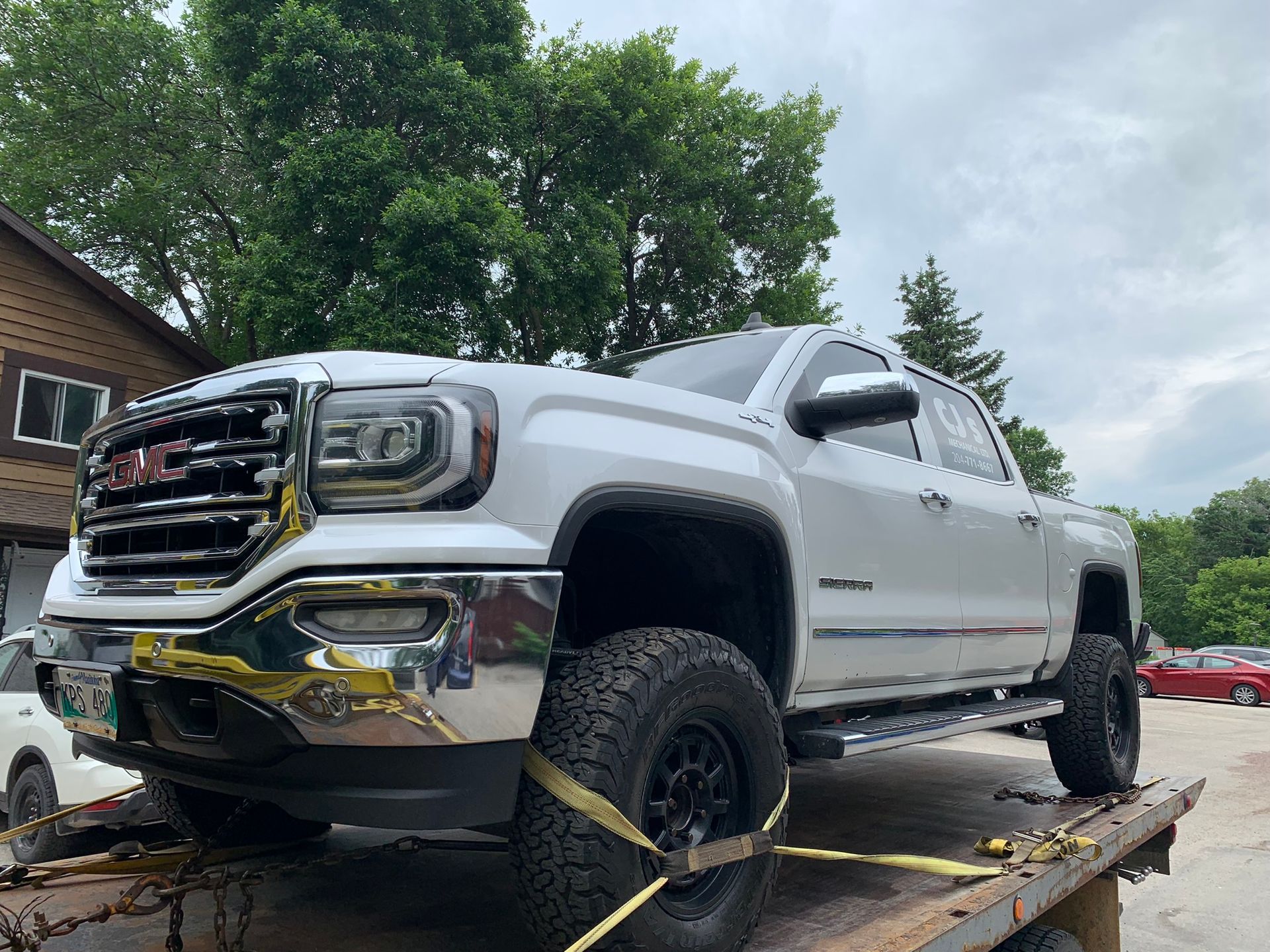 White lifted GMC pickup strapped to a flatbed trailer outdoors under cloudy skies