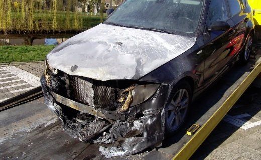 Black car with severe front-end damage, hood crumpled and bumper broken, parked on a tow truck bed.