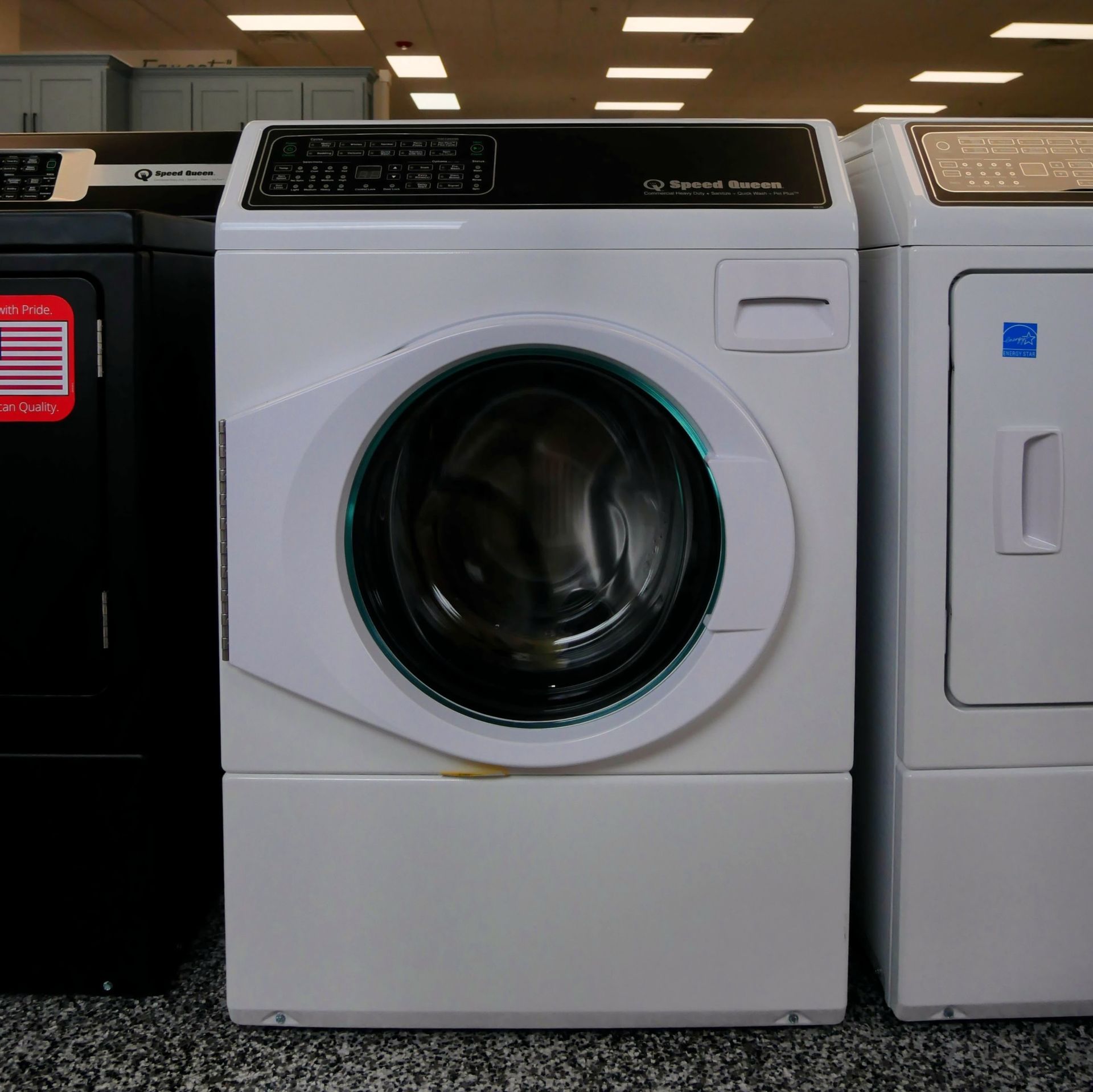 White commercial washing machine with a black control panel, flanked by other appliances.
