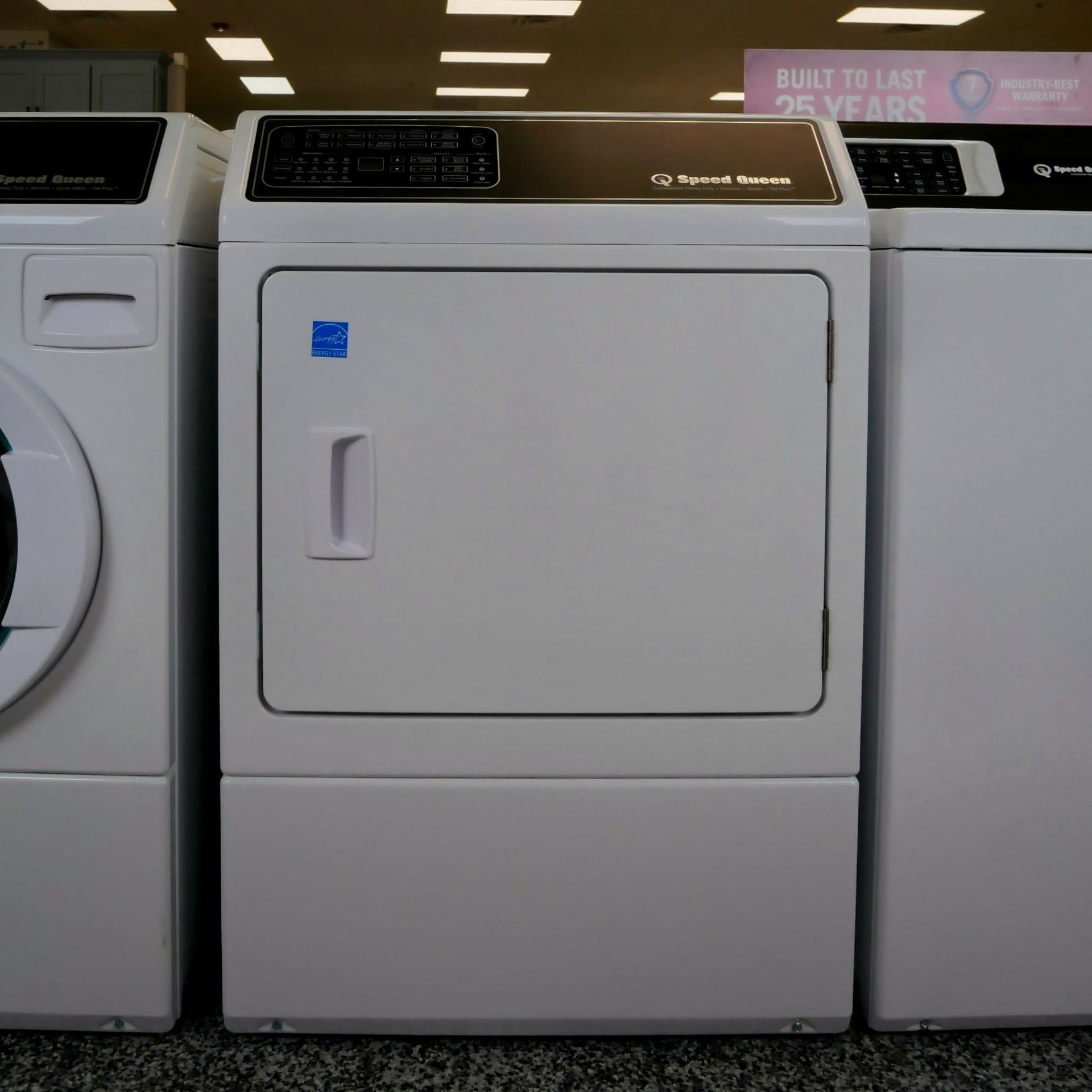 A white commercial clothes dryer flanked by two white washing machines, indoors.