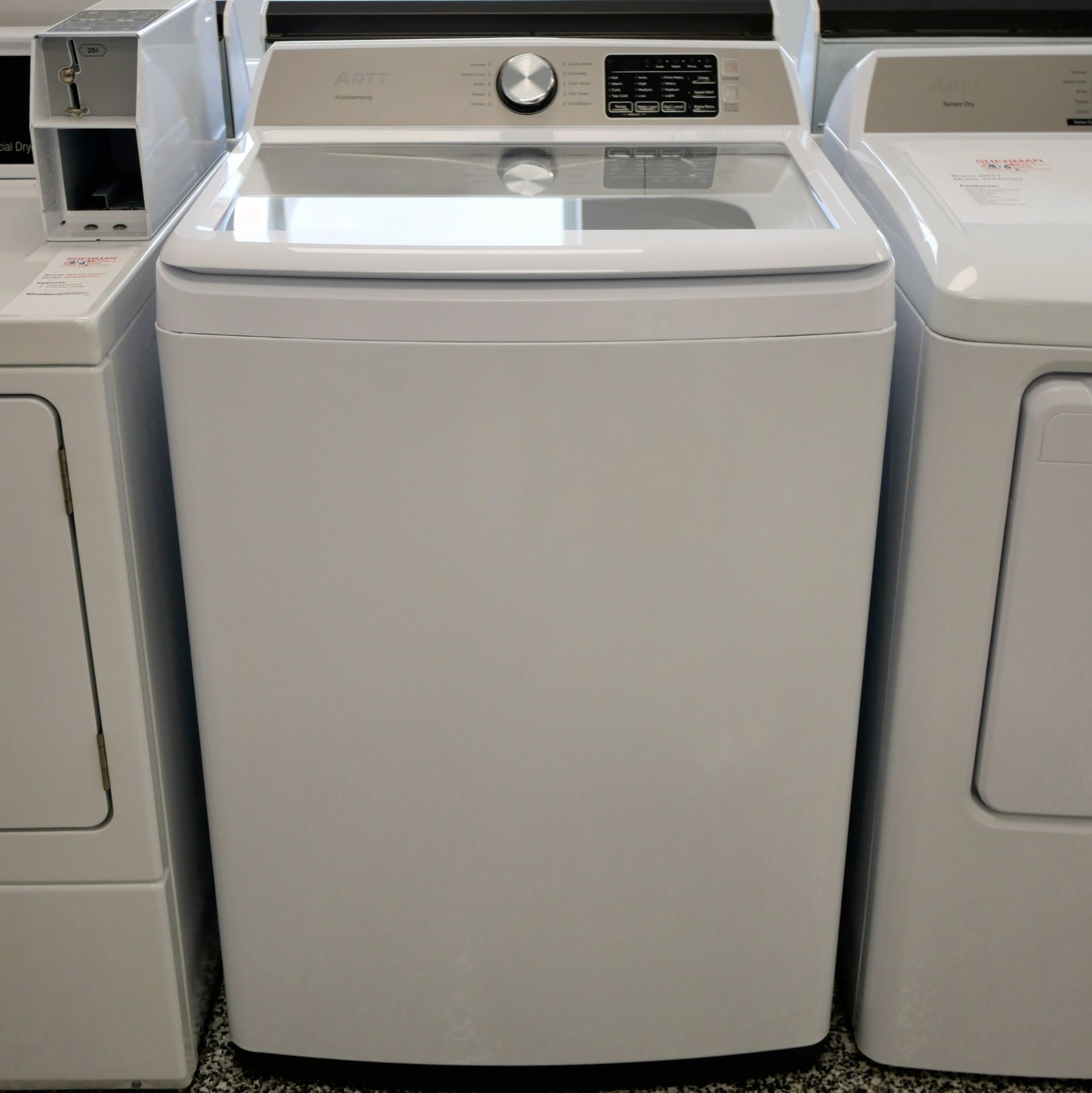 White top-load washing machine with control panel on top; front view, between two other appliances.