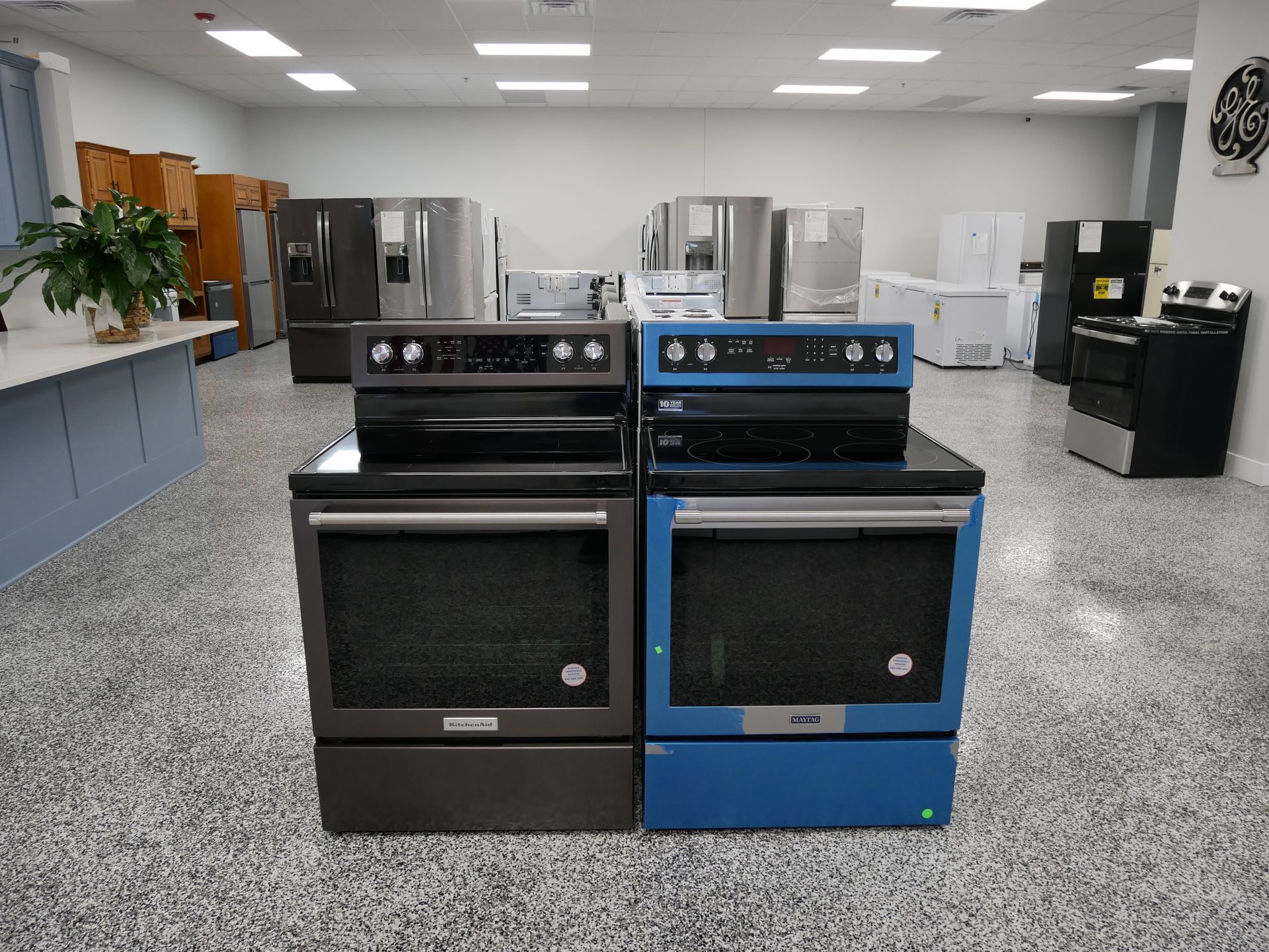 Two electric ovens, one black and one blue, on display in a showroom with other appliances.