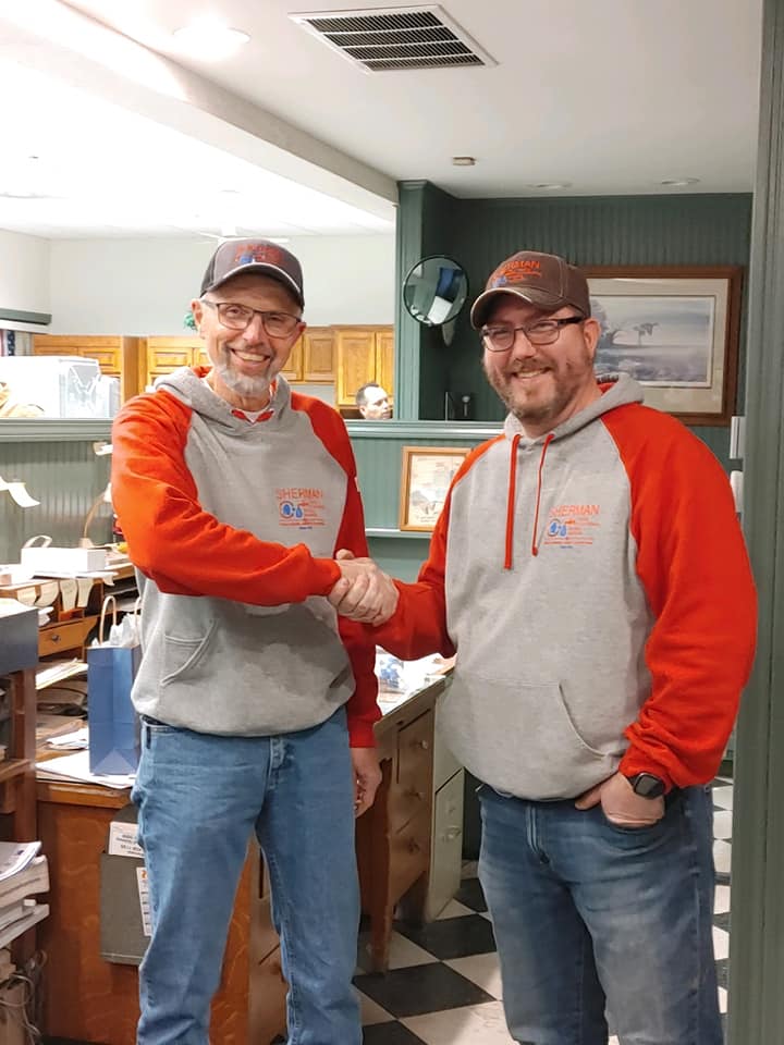 Two men in matching hoodies and hats shaking hands inside a room with wood paneling.