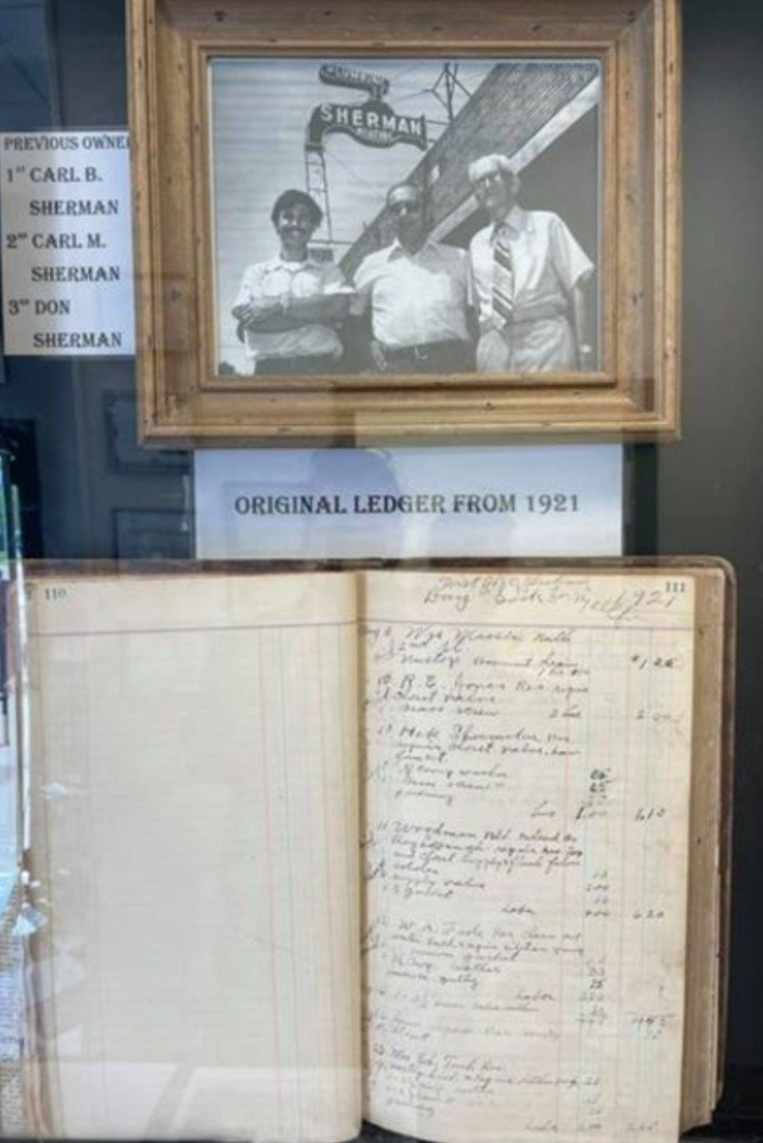 Framed photo of three men above an open ledger from 1925, displayed in a glass case.