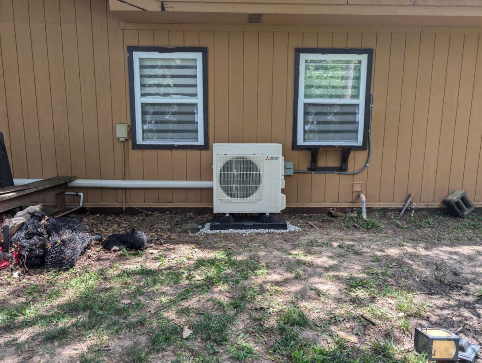 Exterior view of a building with a beige wall, two windows with blinds, and an air conditioning unit on a gravel base.