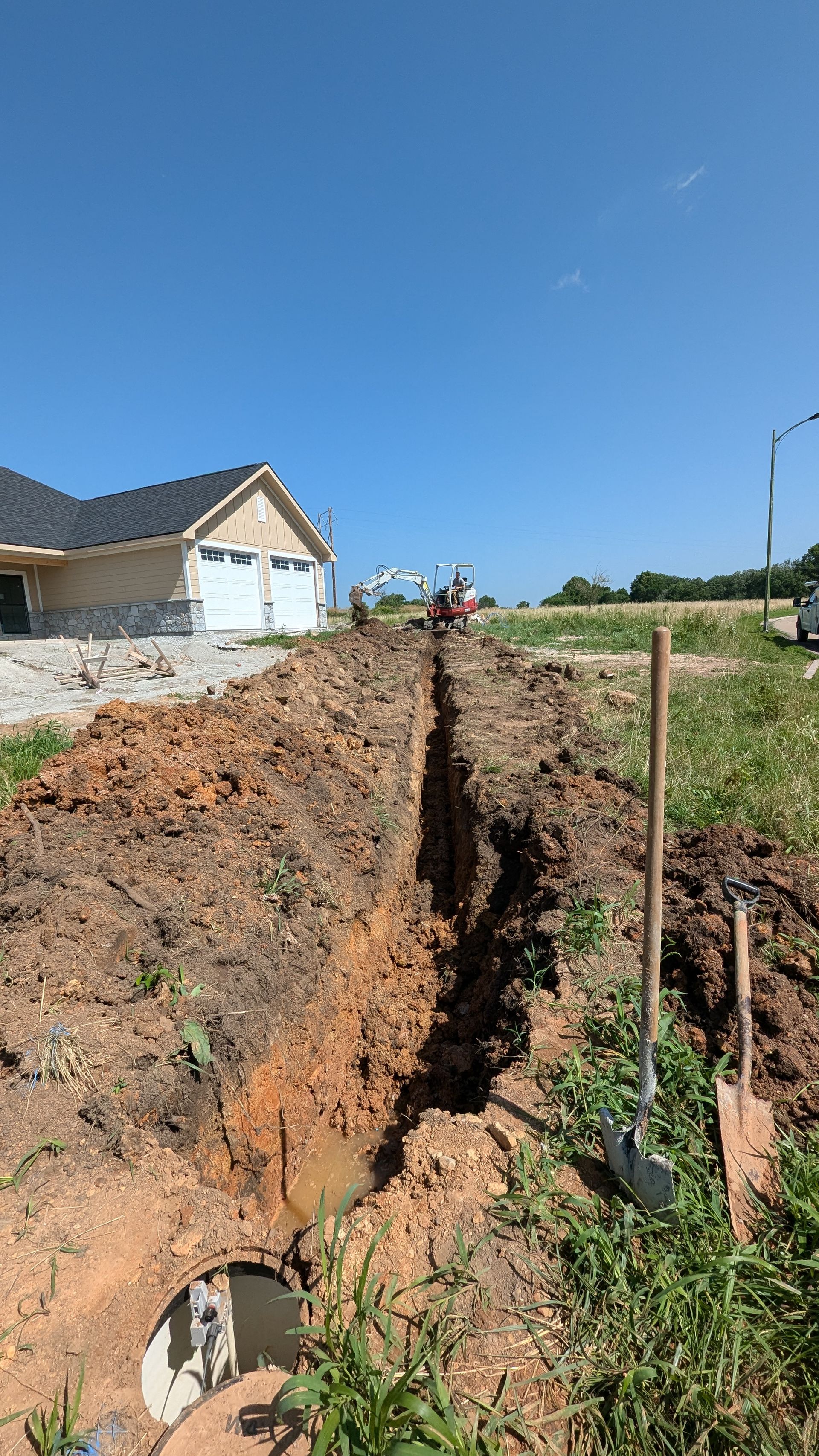 A deep trench being dug in a yard near a house. Shovel leans against the side.