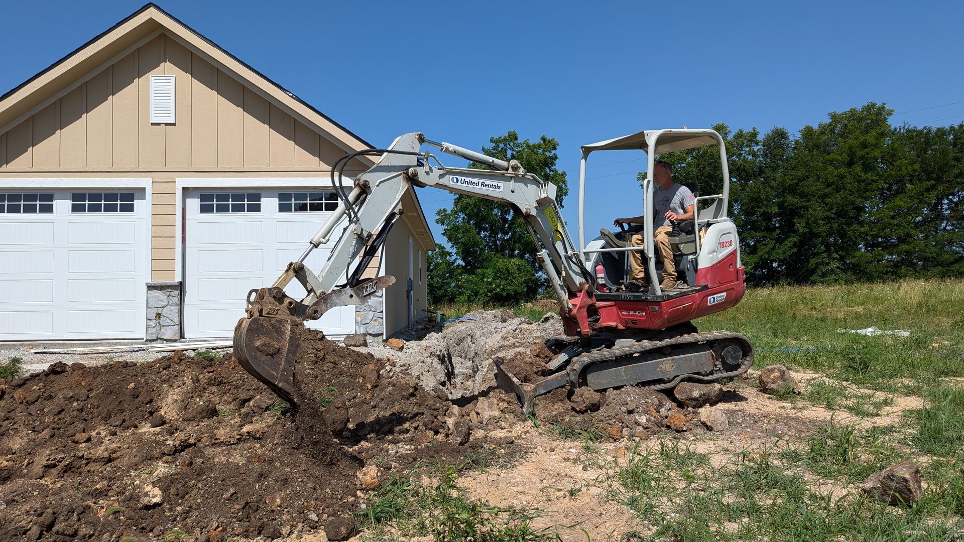 A small excavator is digging a hole in front of a garage.