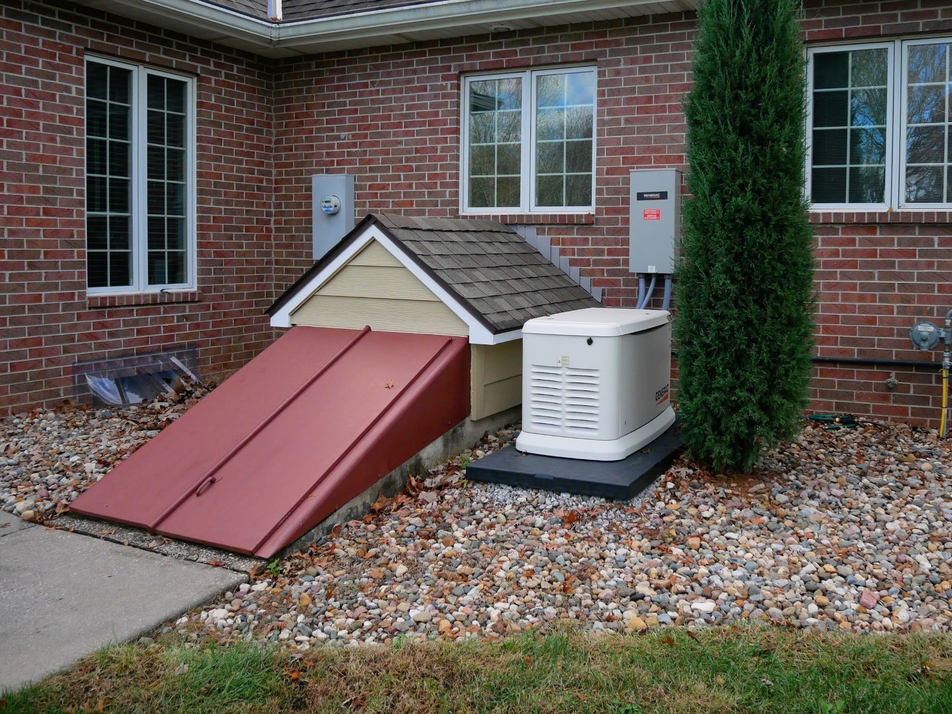 Red-doored basement entrance, generator, electrical boxes near a brick house, next to a tree and gravel.