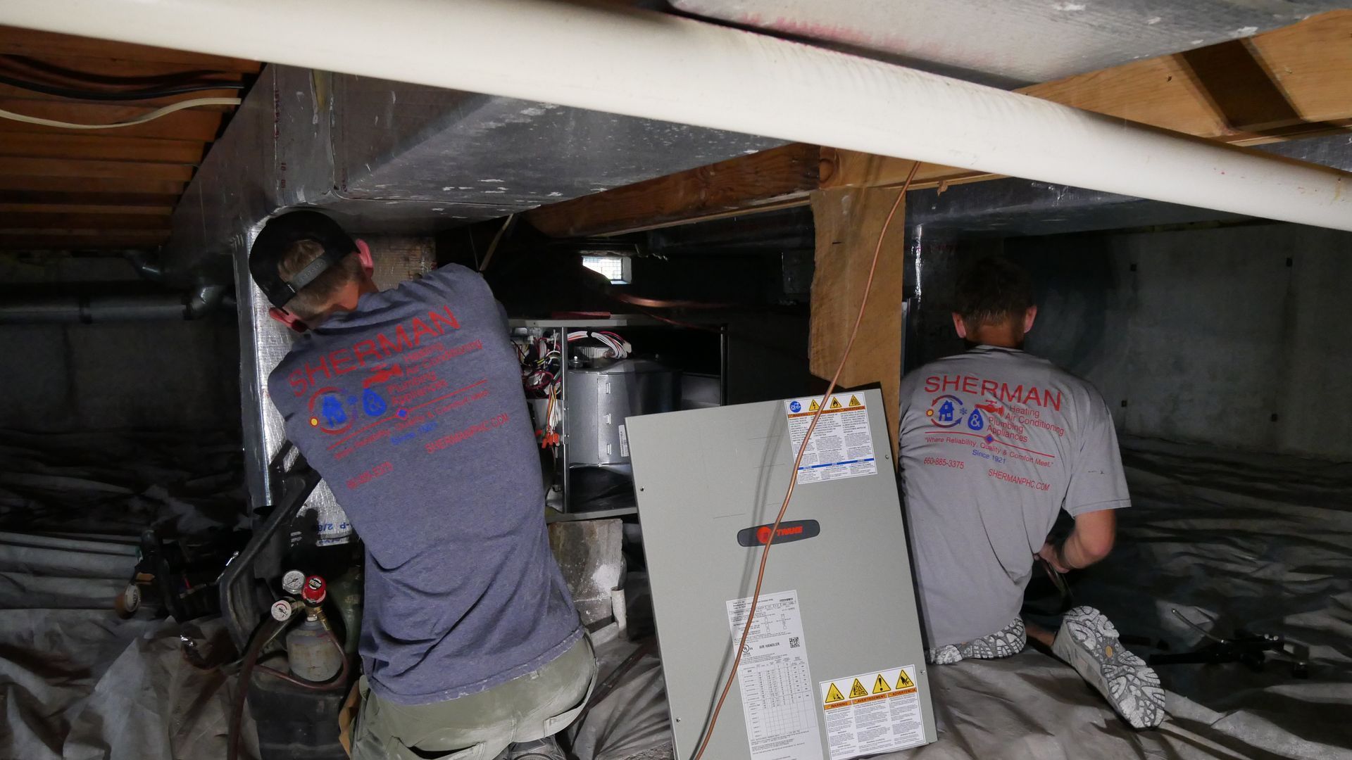 Two men are working on an air conditioner in a basement.