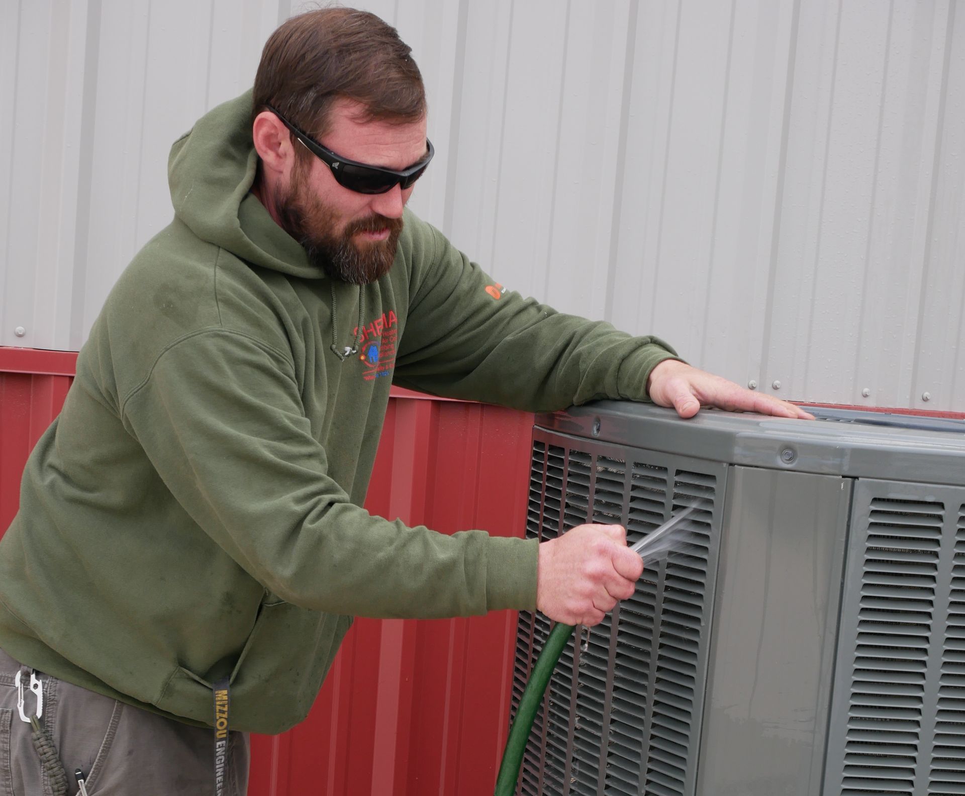 A man in a green hoodie is cleaning an air conditioner with a hose.