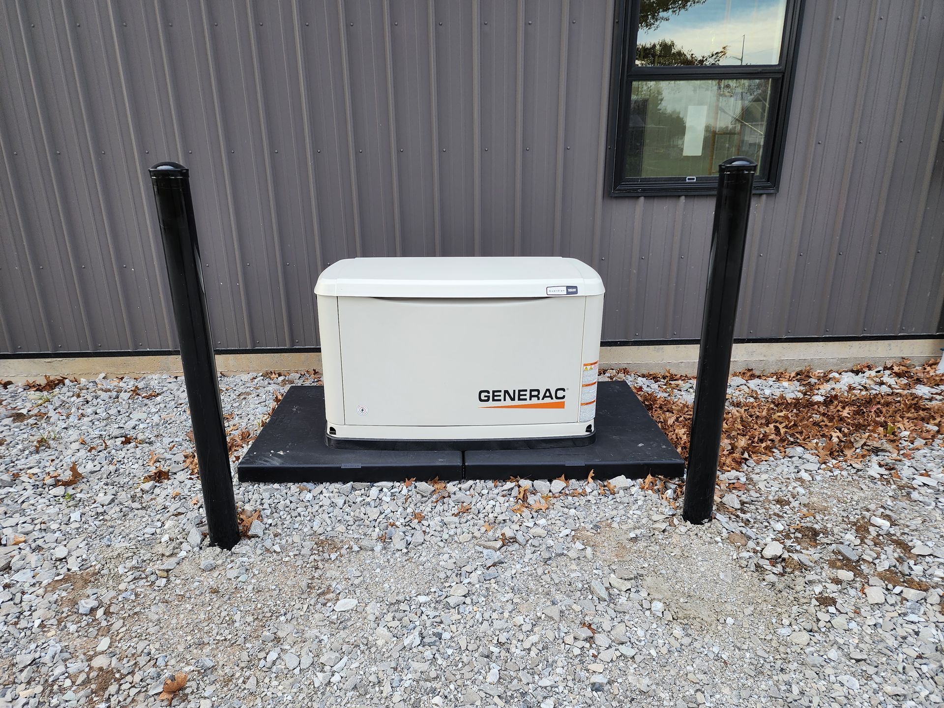 Generator on a black platform between two black posts on gravel, next to a gray building.