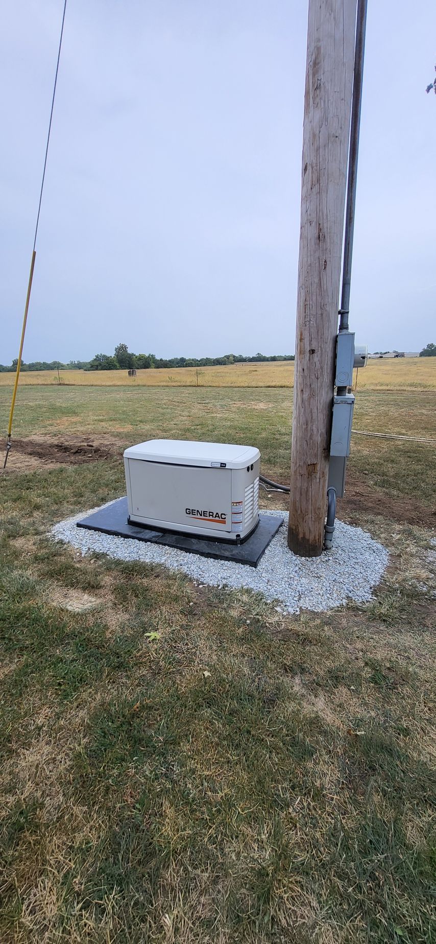 A generator is near a utility pole in a grassy field. The generator sits on a pad surrounded by gravel.