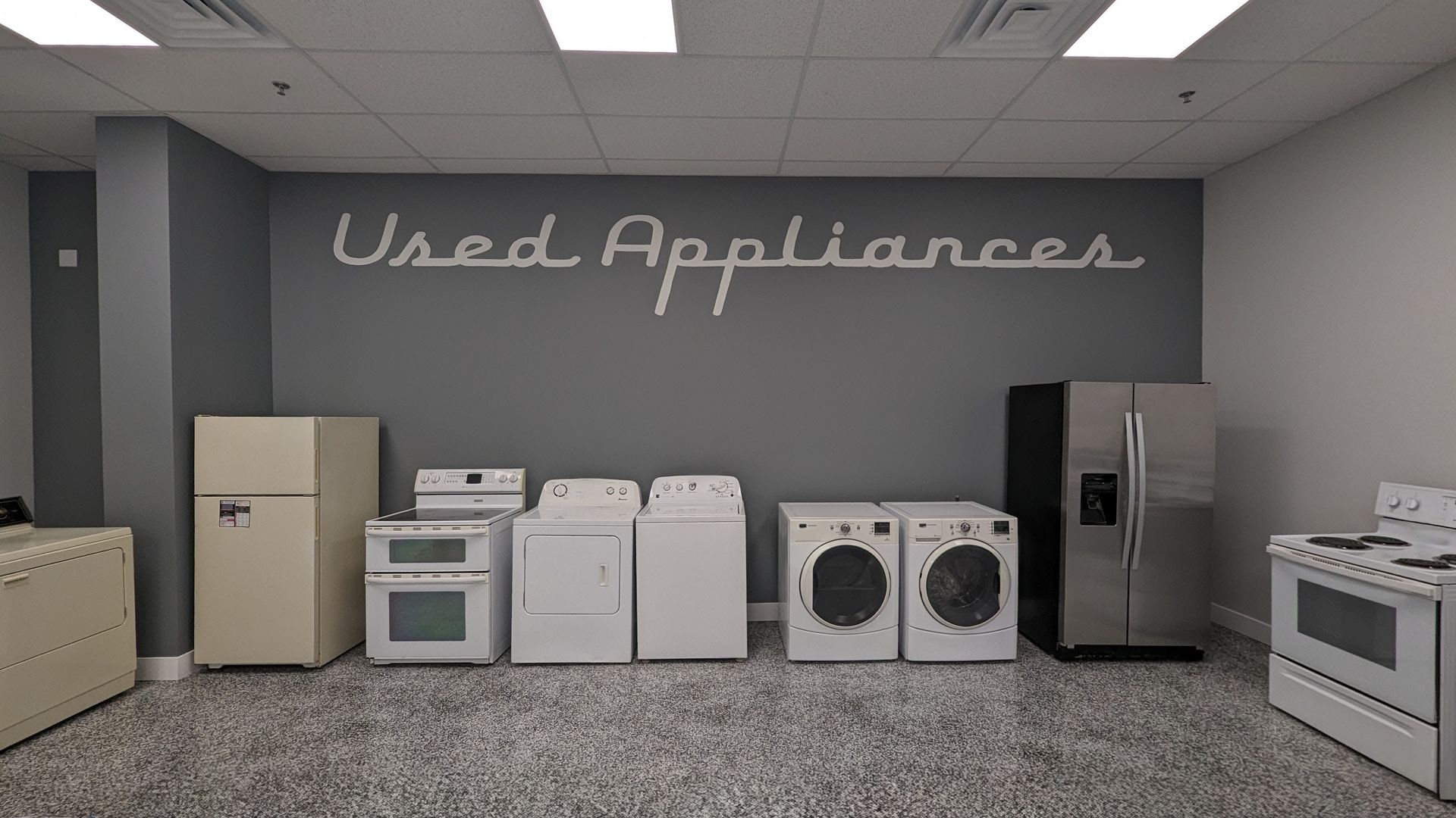 Used appliance store displaying a variety of refrigerators, washers, dryers, and stoves on gray carpet. 