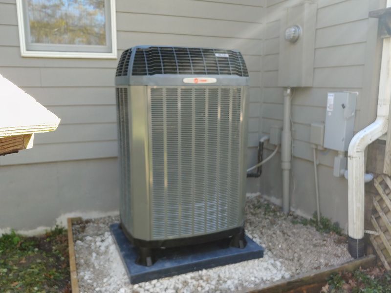 Outdoor Trane air conditioning unit on gravel base next to a house with a window and downspout.