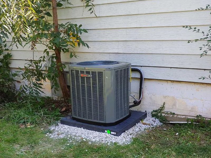 Air conditioner unit next to a light-colored house wall, resting on a black base surrounded by small white stones.