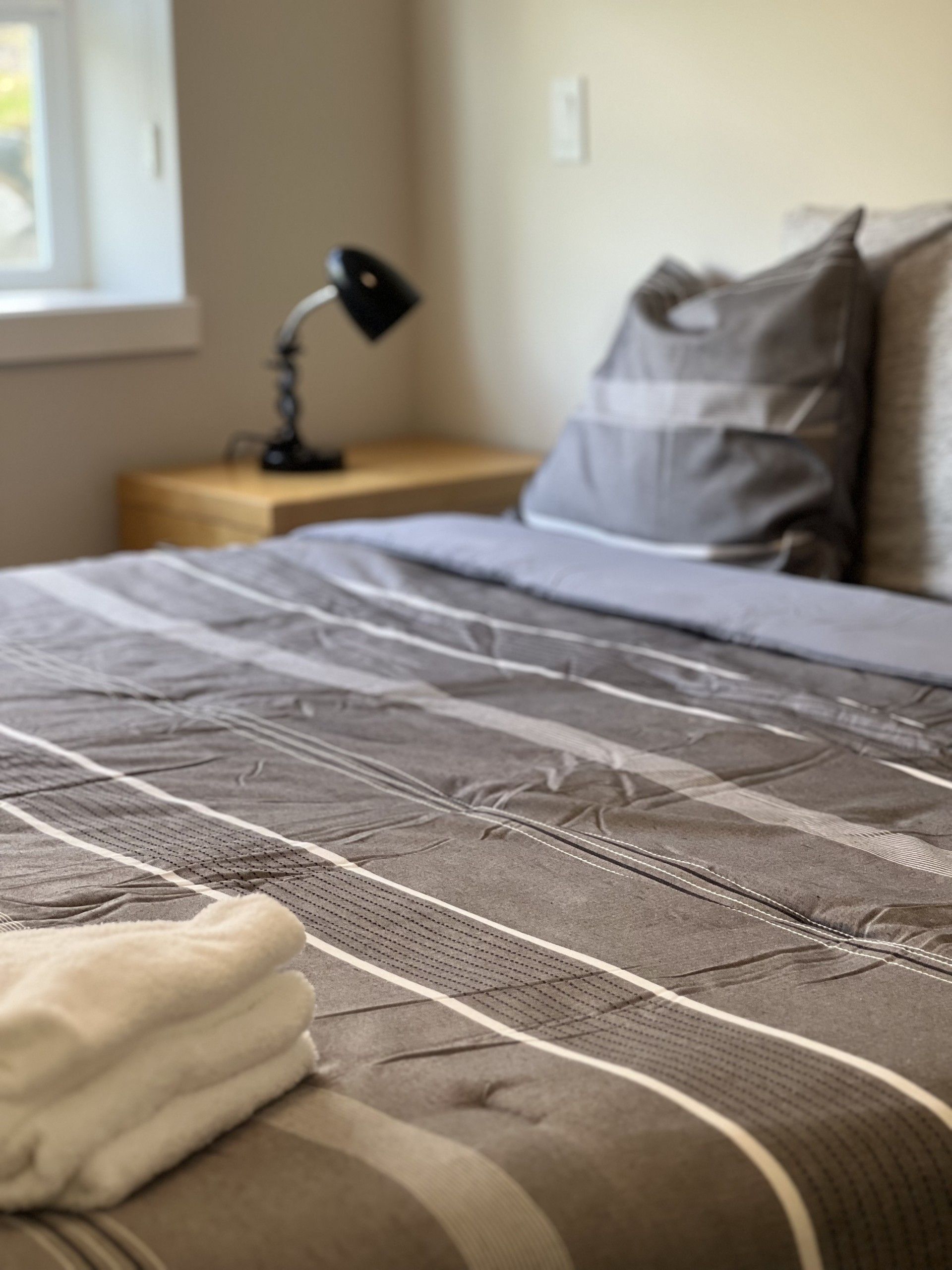 Bedroom with a bed, nightstand, and lamp. Bed has gray and white striped comforter and pillows. Window in background.