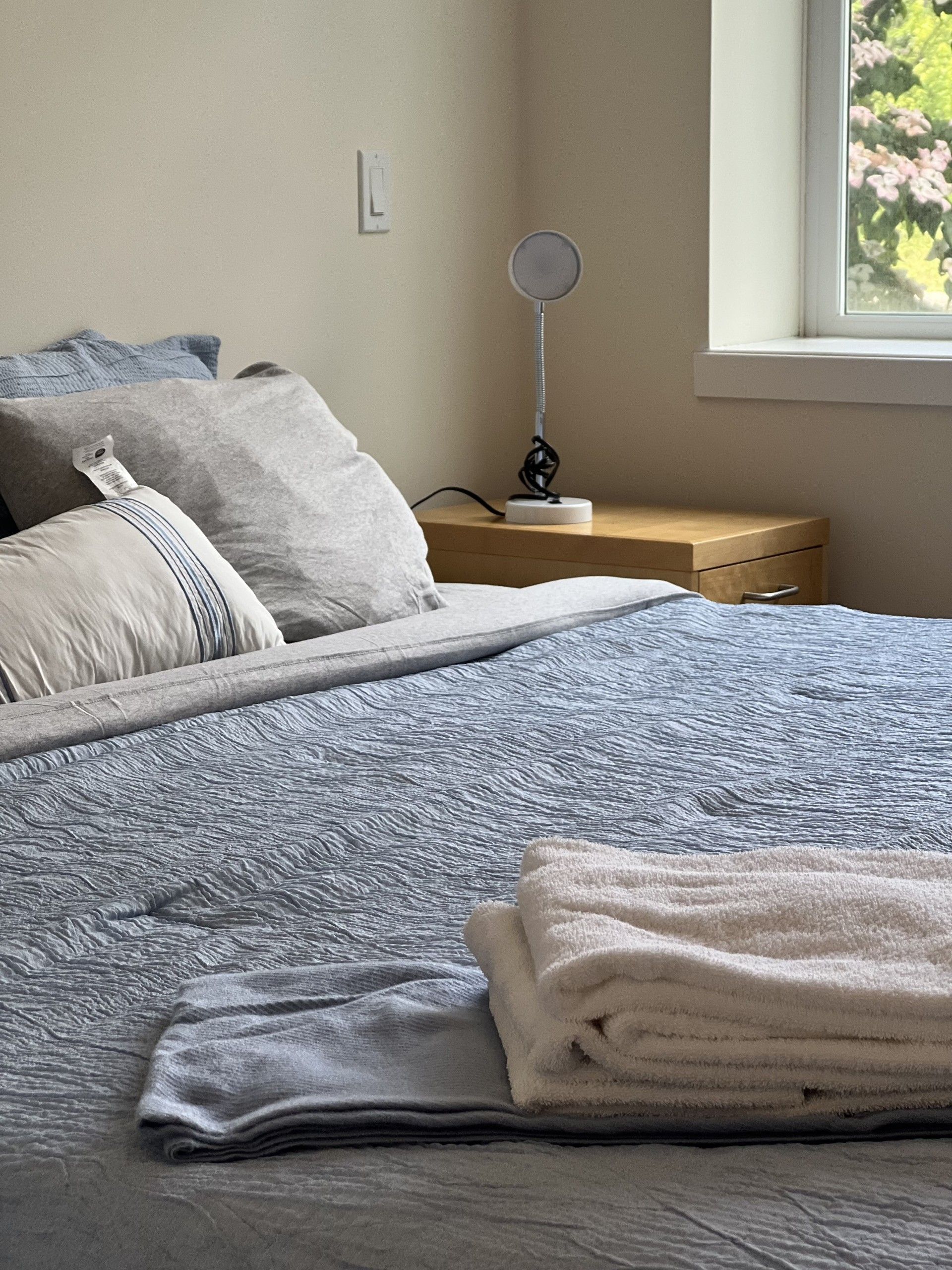 Bedroom with bed covered in blue quilted bedding, pillows, and a stack of towels. A small lamp sits on a wooden nightstand.