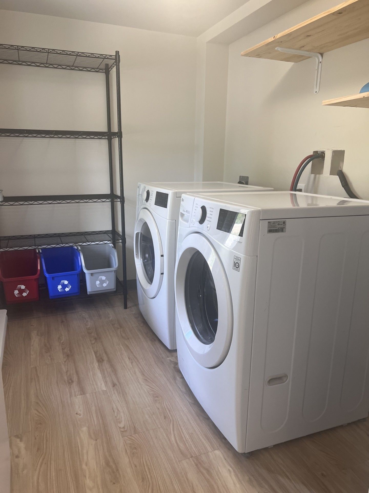 Laundry room with a washer, dryer, recycling bins, shelving, and wooden floor.